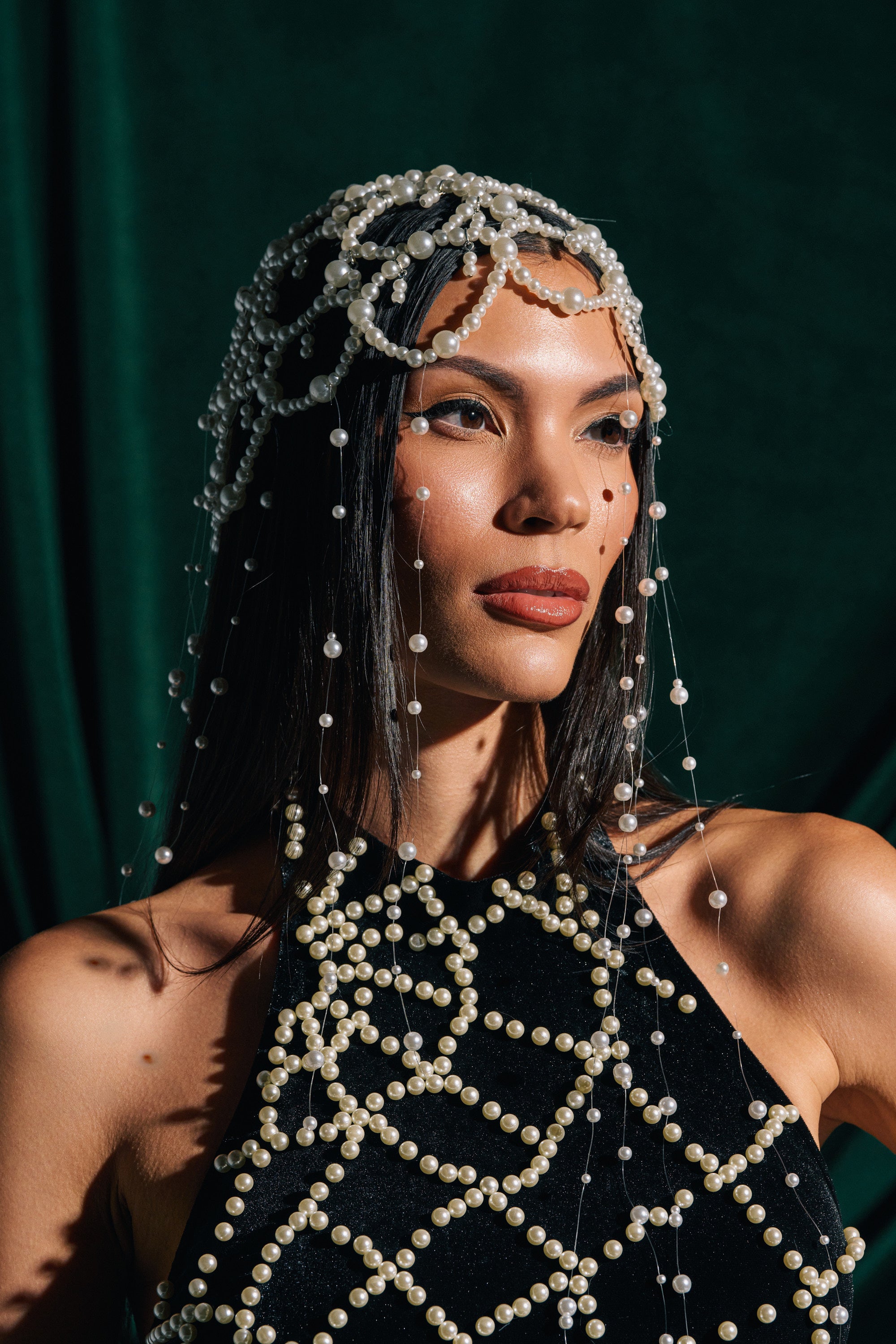 A woman with straight dark hair poses against a dark green background, sunlight illuminating her as she wears the PEARL LOVE HEADPIECE—a delicate pearl veil draped over her head and face—and a black pearl-adorned top.