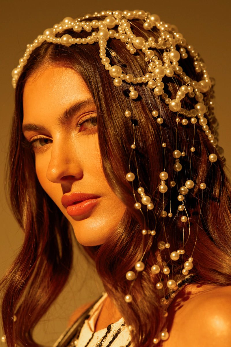 A woman with wavy brown hair wears an elaborate "PEARL LOVE HEADPIECE" featuring cascading strands. She gazes softly at the camera, while warm lighting highlights her features and pearl beads shimmer gently against a muted, warm-hued background.