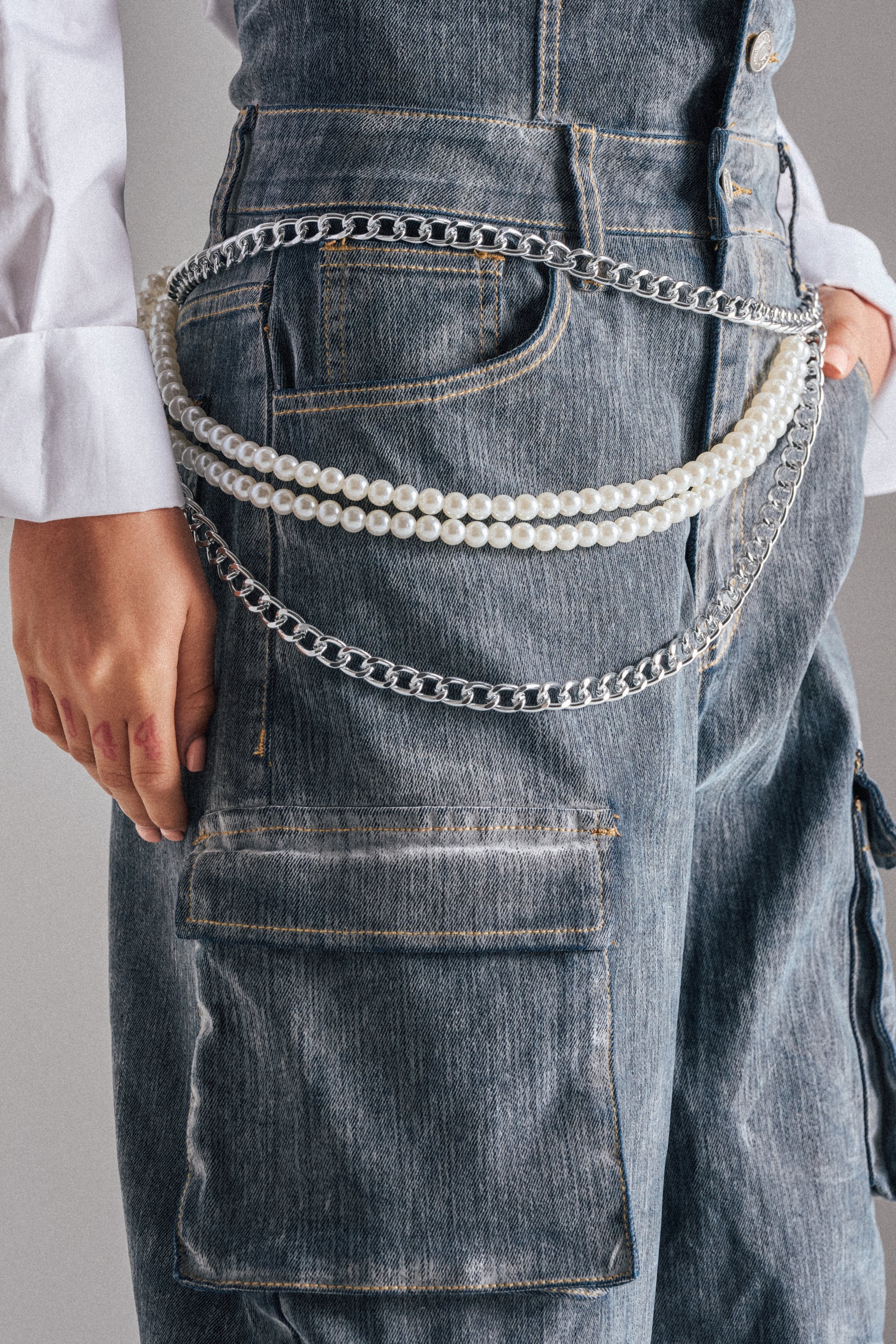 A person wears denim cargo pants with rolled-up cuffs and a white shirt, styled with layered silver chains and the BELT—a pearl chain hip belt—draped statement-style across their hip. 1