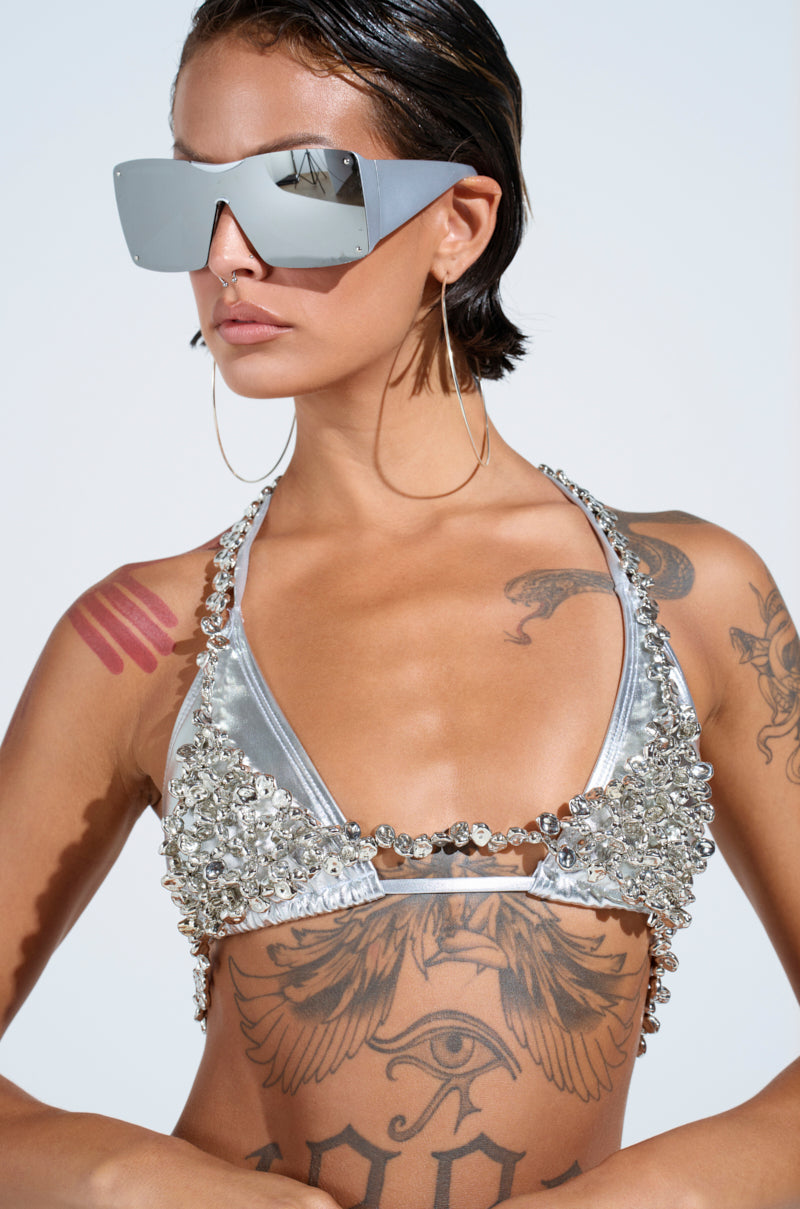 A person wearing large mirrored sunglasses and a shiny, embellished bikini top paired with the KEL BODY CHAIN. They have slicked-back hair, multiple tattoos—including an eye on their chest and script on their arm—against a plain, light background. 1