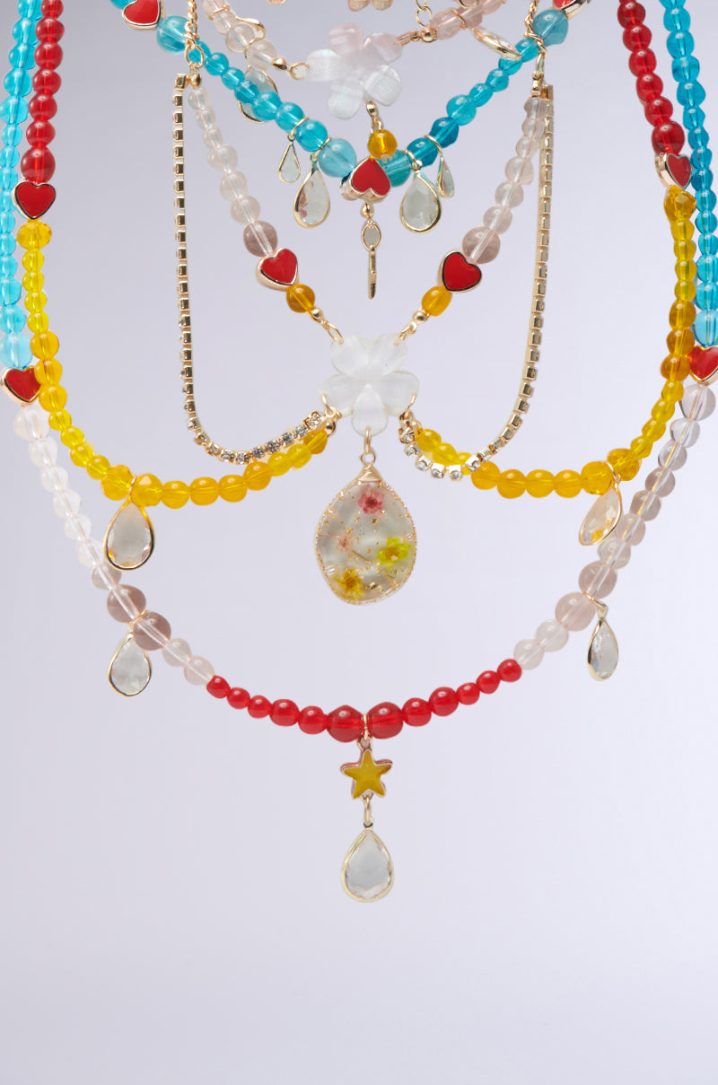 The DIDI LOVE NECKLACE is a vibrant multi-layered piece with multicolored glass beads, featuring heart charms, teardrop pendants, and a central flower-shaped bead in red, blue, yellow, and clear hues against a plain light gray background.