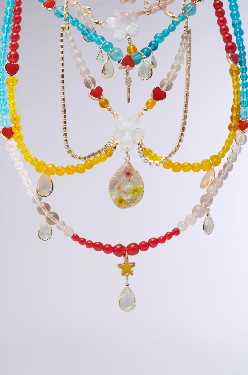 The DIDI LOVE NECKLACE is a vibrant multi-layered piece with multicolored glass beads, featuring heart charms, teardrop pendants, and a central flower-shaped bead in red, blue, yellow, and clear hues against a plain light gray background. 2