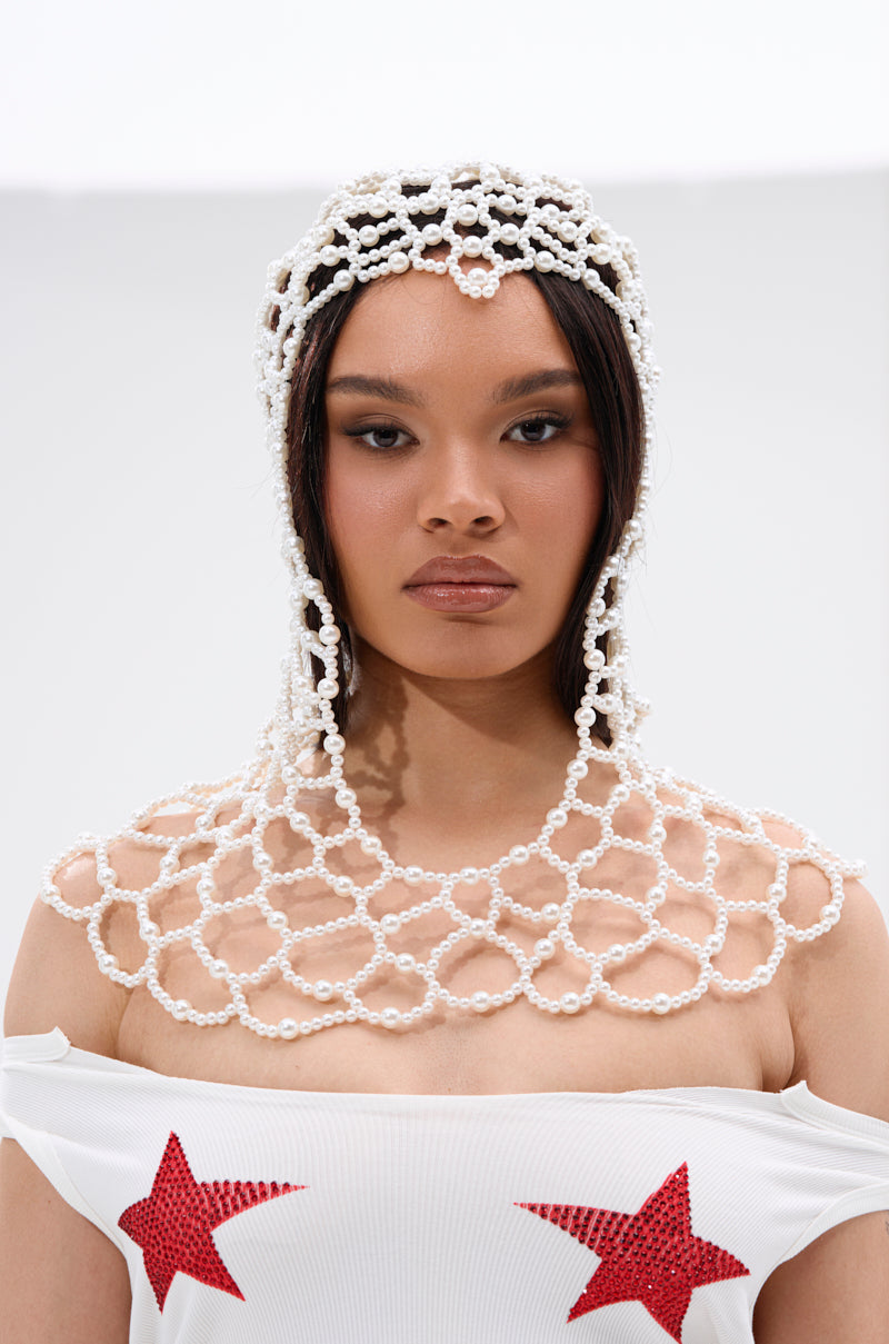 A woman wears the ELEGANT VIBES PEARL HEADPIECE with circular patterns draped over her head and shoulders, paired with a white off-the-shoulder top with red star designs, exuding elegance with her neutral expression.
