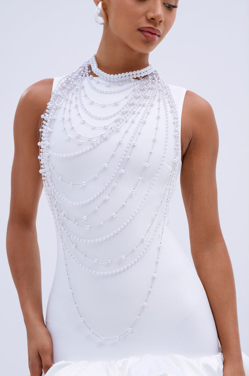 A woman in a sleeveless white dress with a flowing silhouette poses against a light background, wearing the IM TOO PRETTY NECKLACE—a long, layered statement piece featuring faux pearls and crystals. 0