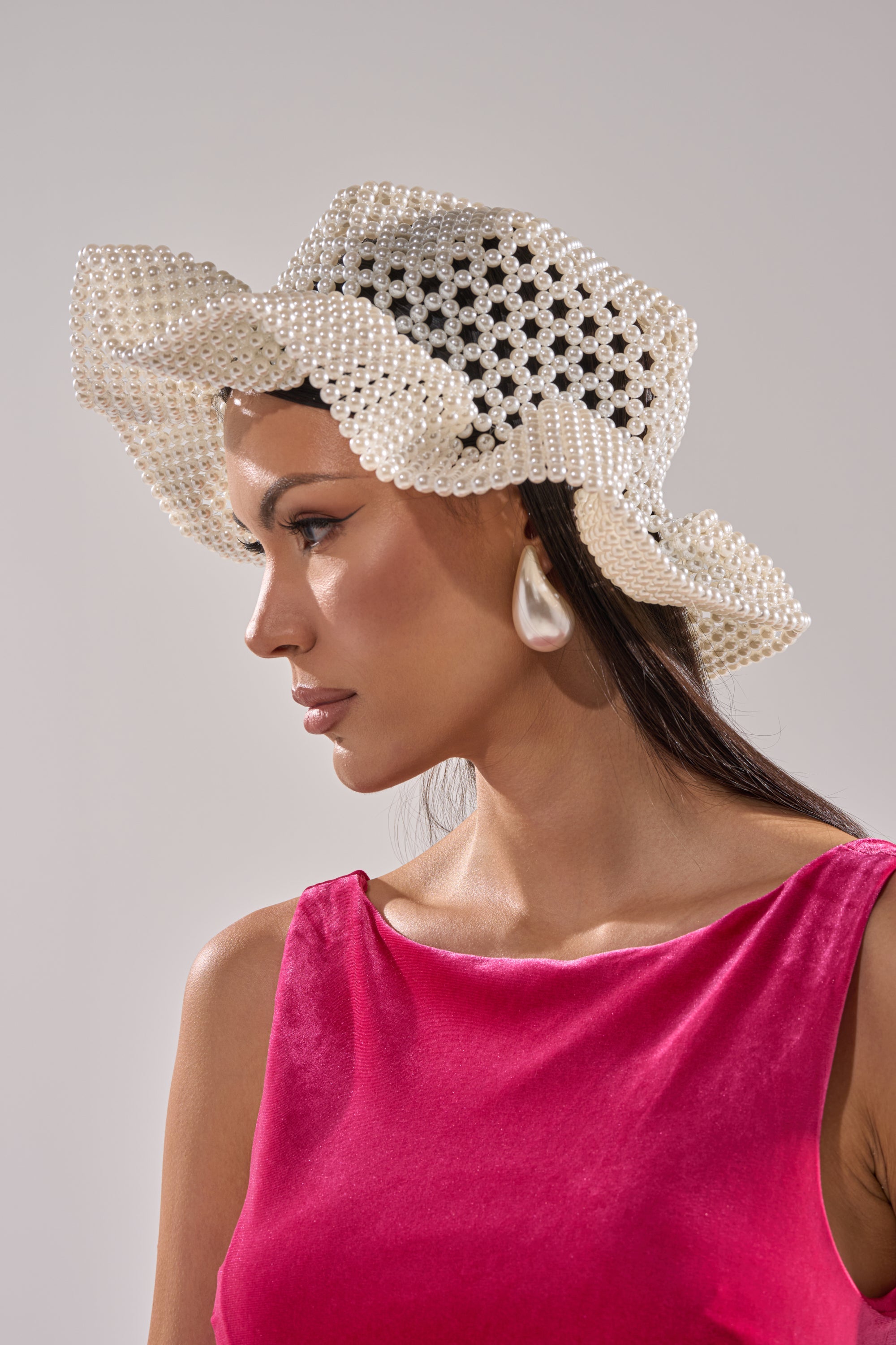 Woman in a sleeveless pink velvet top wears the PEARL DREAMS BUCKET HAT and pearl drop earrings, looking left against a neutral background. 0