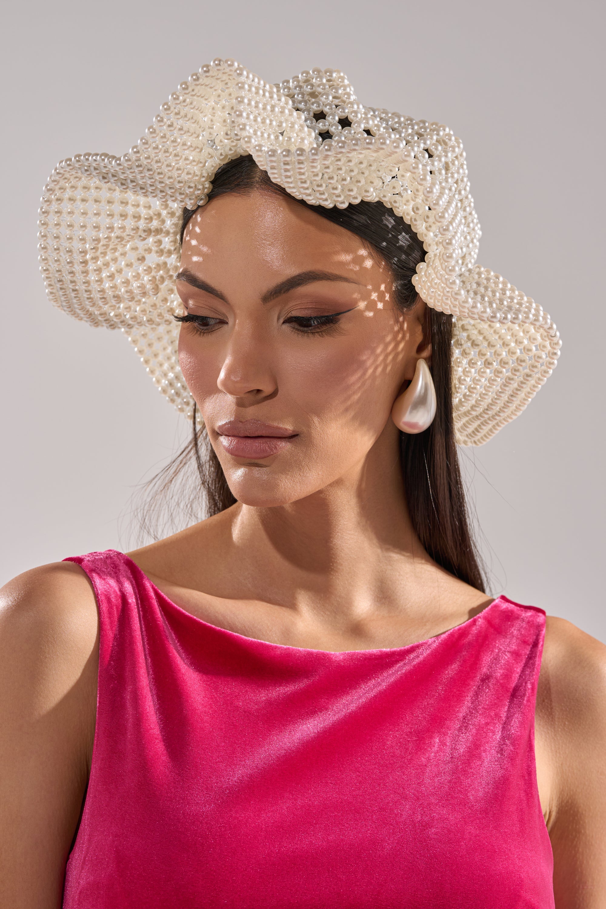 A woman in a pink velvet sleeveless top wears the PEARL DREAMS BUCKET HAT and teardrop earrings. Soft light casts patterned shadows on her serene face as she looks down, set against a neutral background. 1