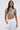 A woman with long brown hair confidently poses against a plain background, wearing the CLEOPATRA RHINESTONE CHAIN TOP—with a pearl collar neckline and faux pearls—paired with white high-waisted pants and one hand in her pocket.