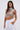 A woman with long straight brown hair wears the CLEOPATRA RHINESTONE CHAIN TOP featuring a pearl collar neckline, paired with white zip-pocket pants. She places one hand in her pocket and carries a metallic purse.