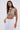A woman with straight brown hair wears the CLEOPATRA RHINESTONE CHAIN TOP with a pearl collar neckline and faux pearls, paired with high-waisted white pants. She stands against a plain white background carrying a silver bag.