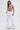 A woman models the CLEOPATRA RHINESTONE CHAIN TOP, featuring a pearl collar neckline, paired with high-waisted white wide-leg pants with textured hems, standing against a plain white background.
