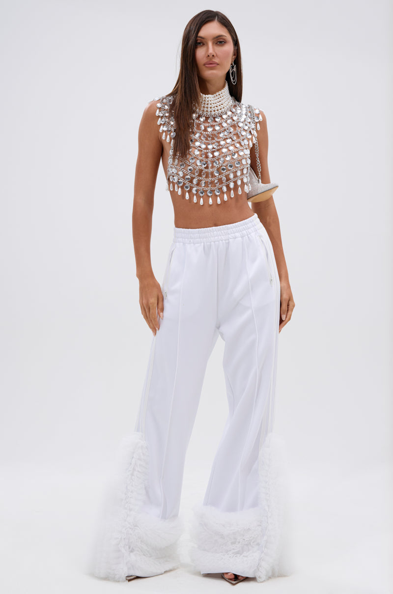 A woman poses against a white backdrop in the CLEOPATRA RHINESTONE CHAIN TOP featuring a pearl collar neckline, paired with wide-legged white pants trimmed with fluffy hems and bold statement earrings for a striking, futuristic look.