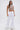 A woman poses against a white backdrop in the CLEOPATRA RHINESTONE CHAIN TOP featuring a pearl collar neckline, paired with wide-legged white pants trimmed with fluffy hems and bold statement earrings for a striking, futuristic look.