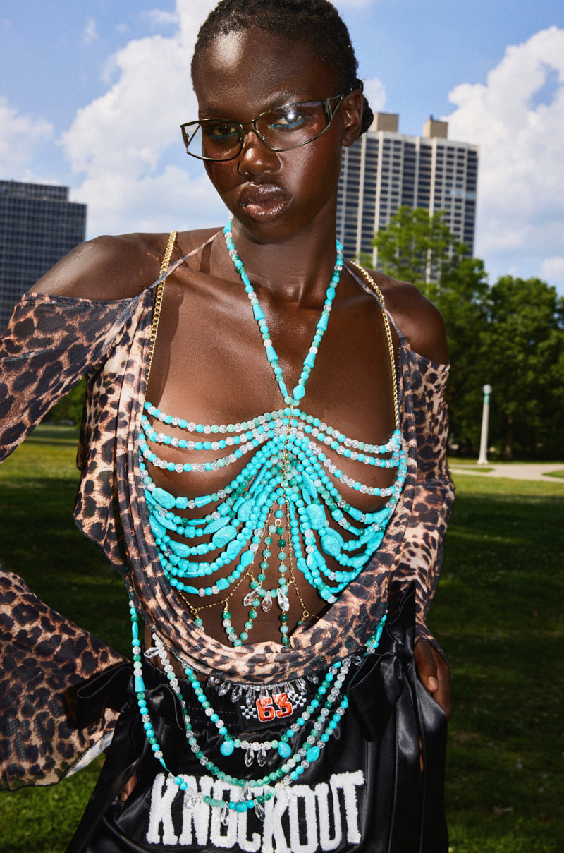 A person with dark skin and glasses poses outdoors in a leopard-print top with cut-out shoulders, layered turquoise bead necklaces, the BEACH GIRLY BRALETTE, and holds a black "KNOCKOUT" bag against tall city buildings.