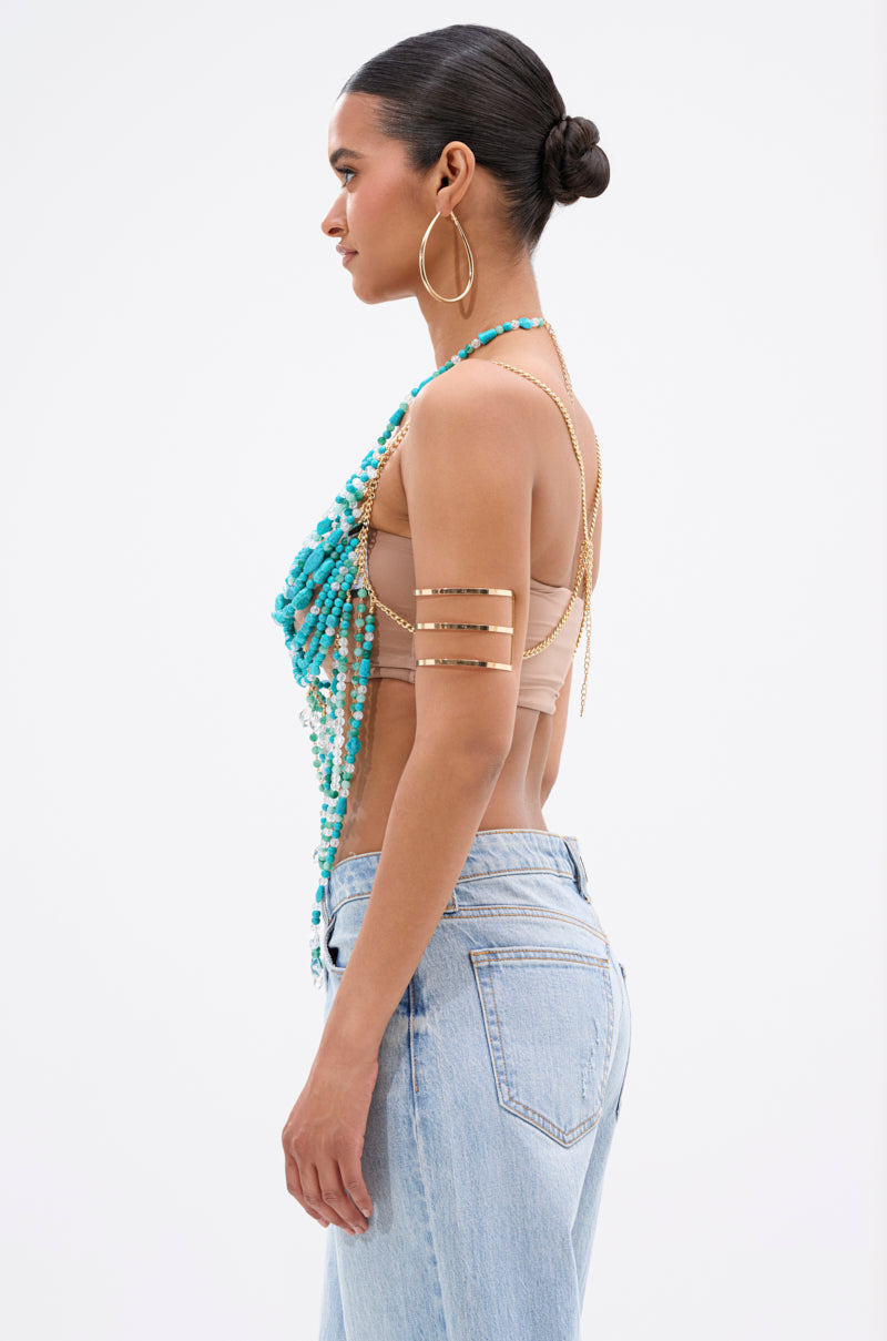 A woman in profile wears the BEACH GIRLY BRALETTE with light blue jeans, turquoise beads, large hoops, and gold cuffs. Her hair is in a bun against a white background, showcasing a chic summer style.
