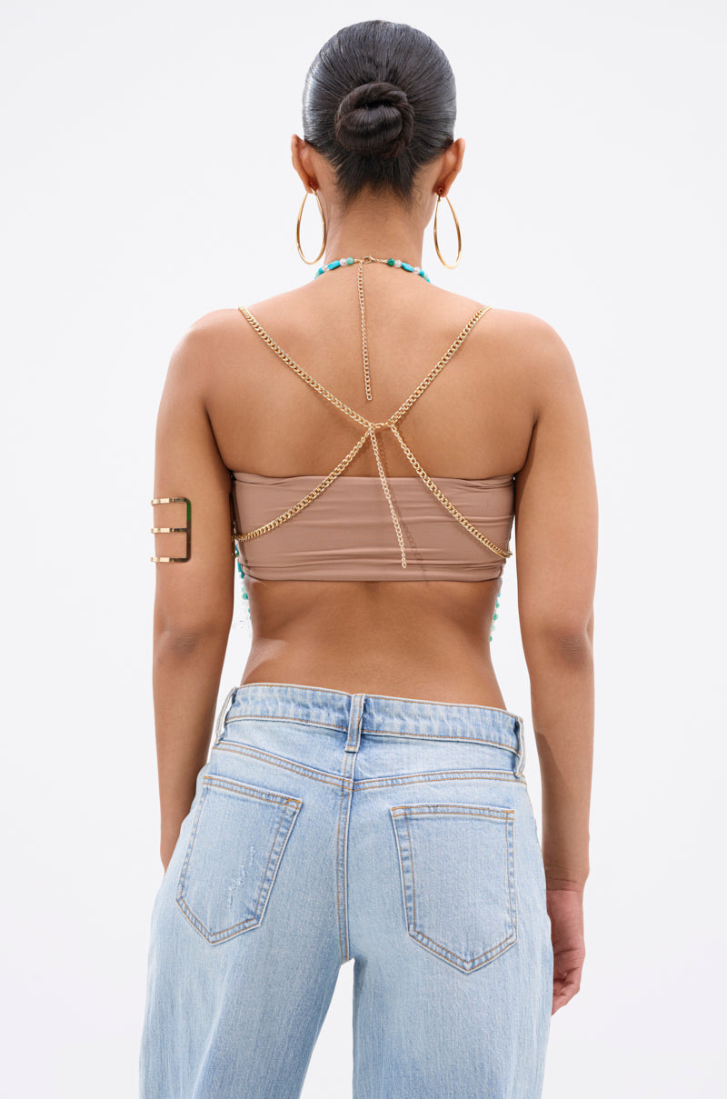 A person with hair in a bun, gold hoop earrings, the BEACH GIRLY BRALETTE strappy top with gold chains, light blue jeans, and an arm accessory stands facing away against a plain white background.