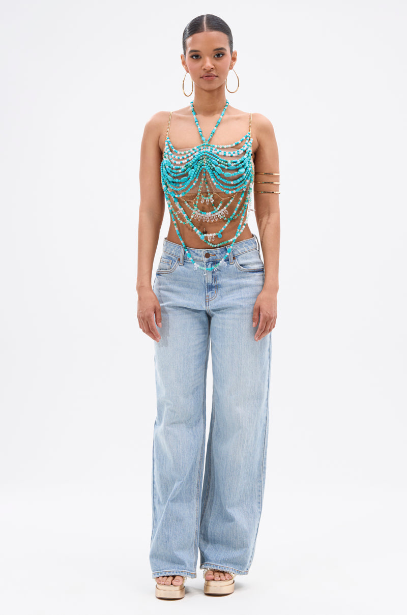 A woman faces forward, wearing hoop earrings, the BEACH GIRLY BRALETTE—a turquoise beaded halter top with draped strands—light blue wide-leg jeans, platform sandals, an arm cuff. Her hair is pulled back and she shows a neutral expression.