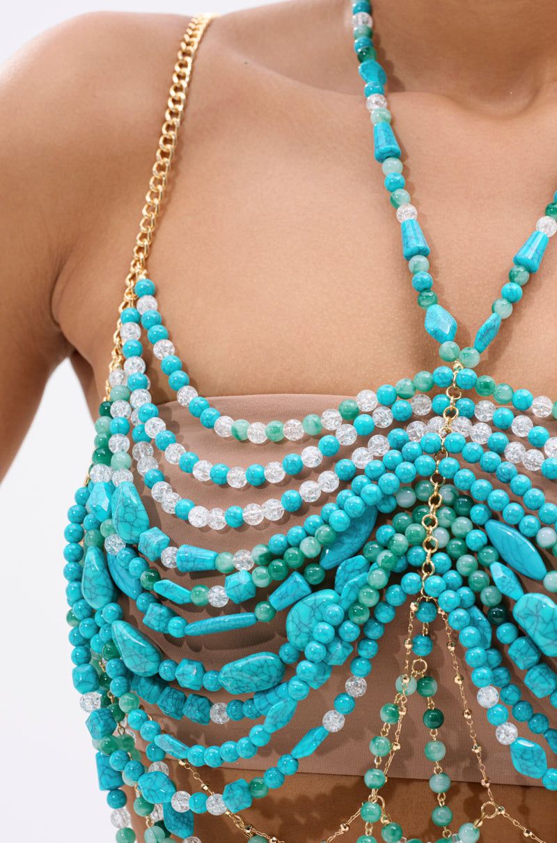 A close-up of a person wearing the BEACH GIRLY BRALETTE, featuring layered turquoise beads and gold chains over sheer nude fabric, with the turquoise beads draped in multiple strands across the chest.