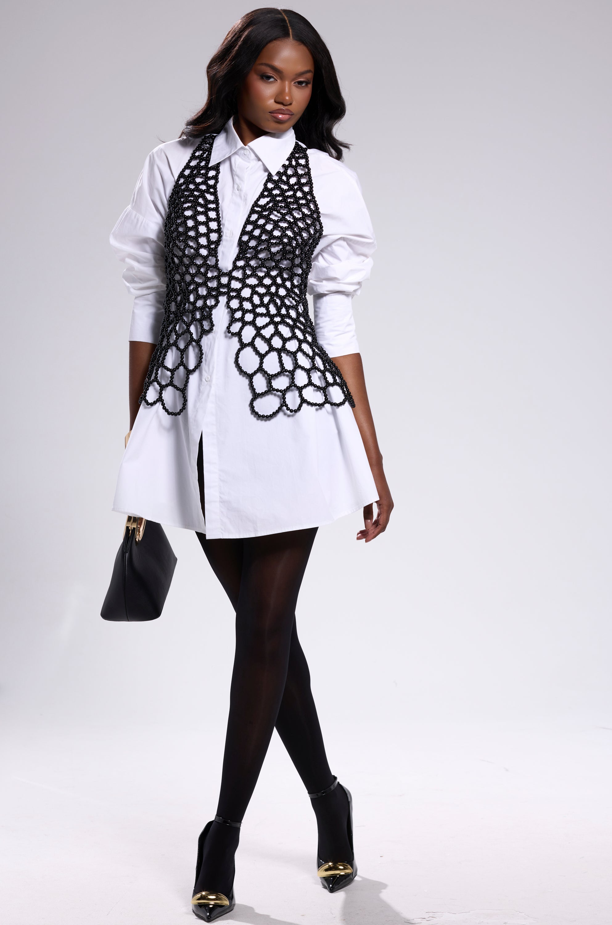 A woman poses in a long white shirt dress, layered with the GETTIN HOT IN HERE BEADED TOP IN BLACK, black tights, ankle boots with gold toes, and a small black handbag against a plain light background. 9