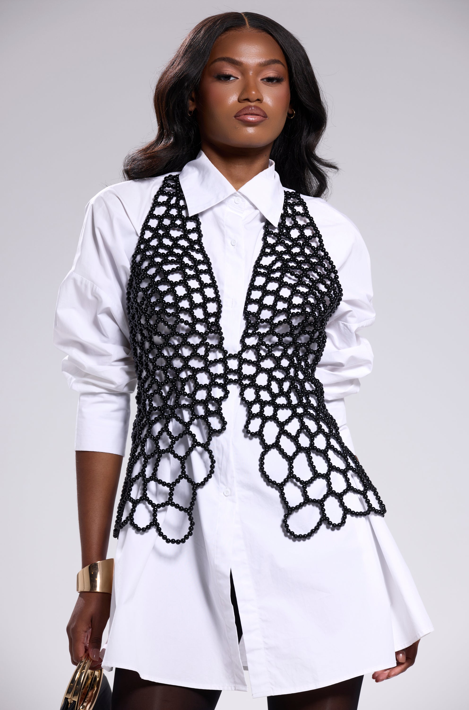 A woman wears a white shirt dress layered with the AKIRA Label GETTIN HOT IN HERE BEADED TOP IN BLACK. She has long wavy hair, gold jewelry, holds a clutch purse, and stands against a plain light gray background. 3