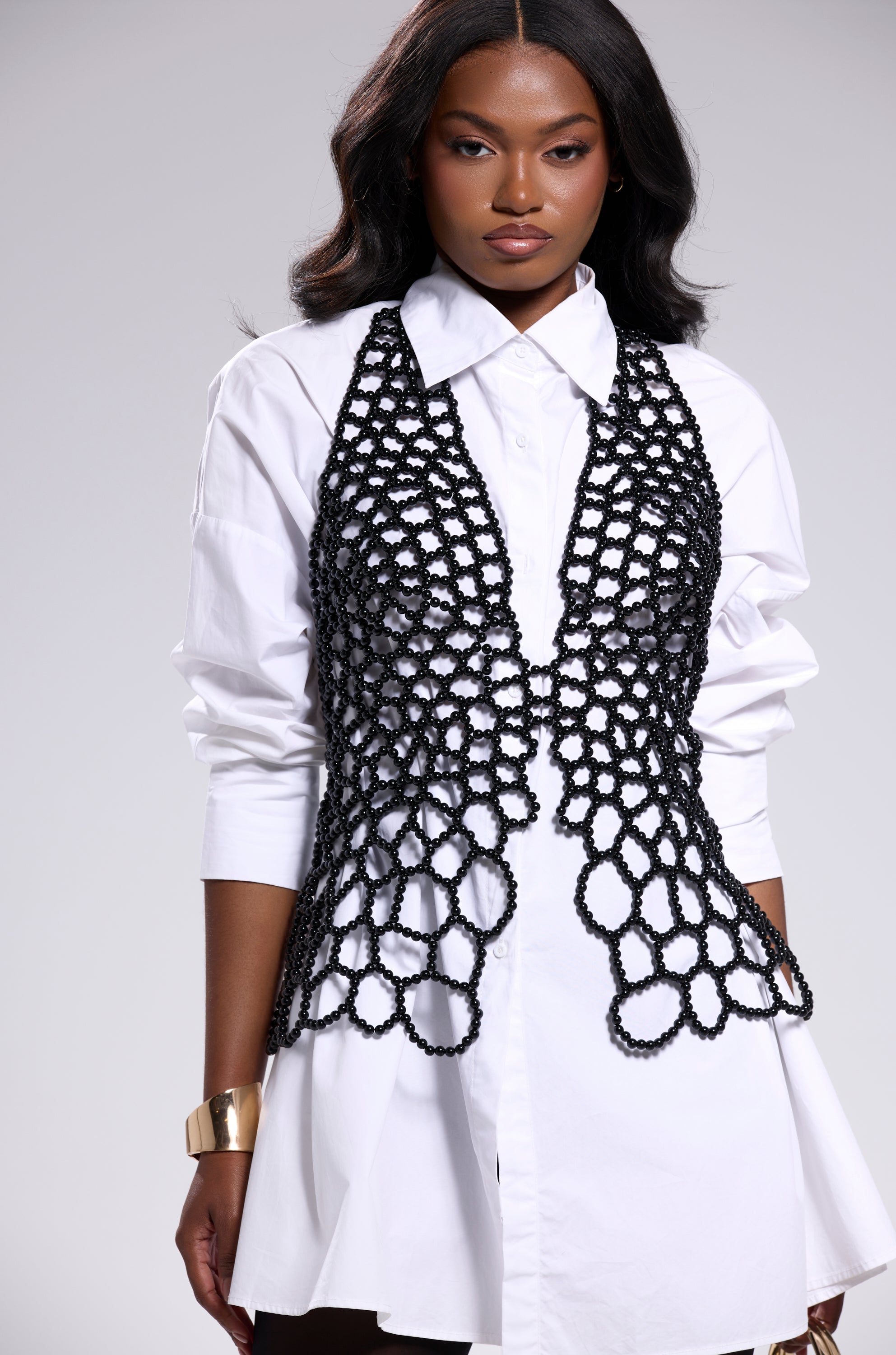 A woman with long wavy hair stands against a light gray background, wearing a white button-down dress layered with the GETTIN HOT IN HERE BEADED TOP IN BLACK and accessorized with a gold cuff bracelet. 5