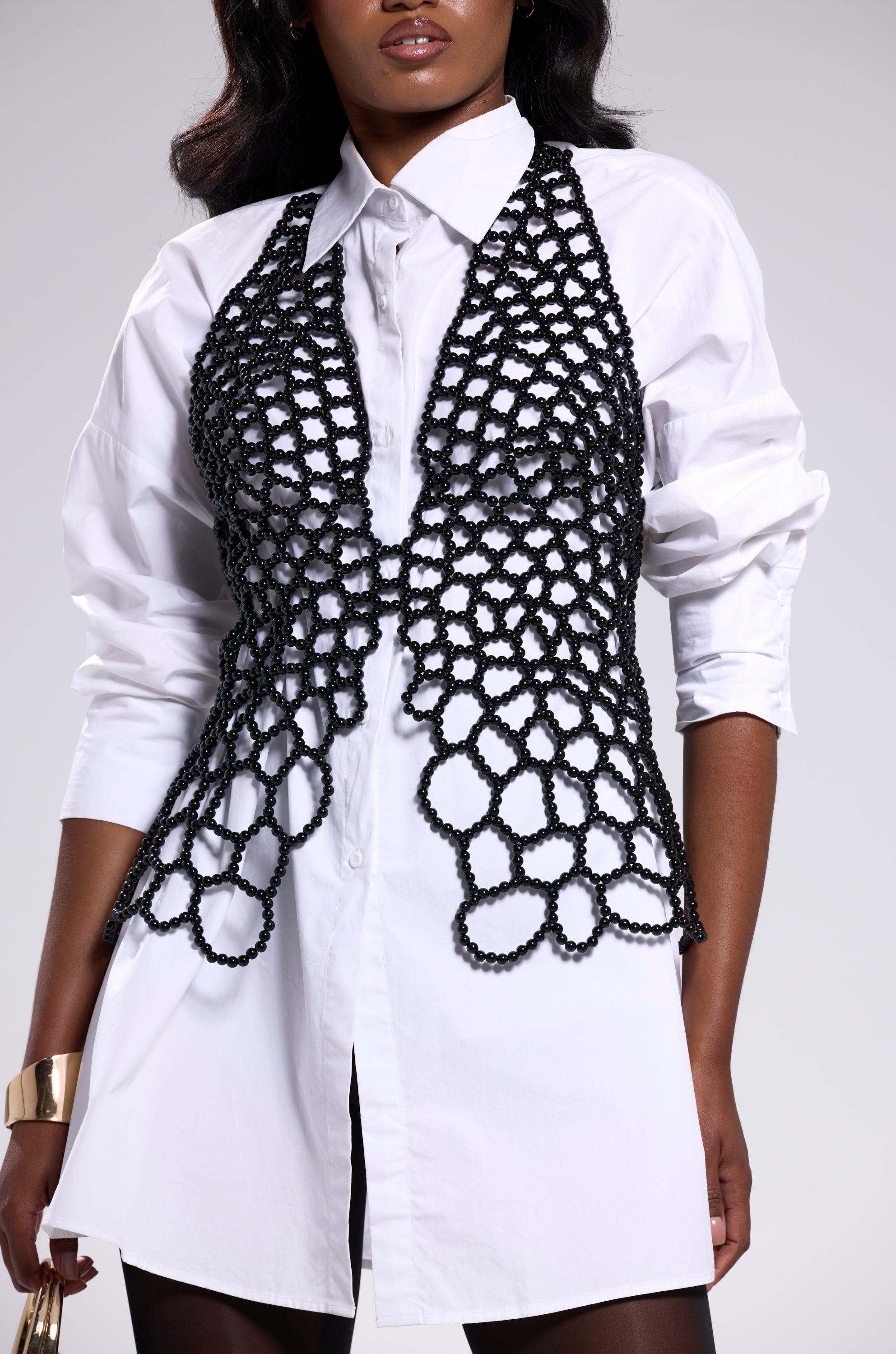 A woman styles the GETTIN HOT IN HERE BEADED TOP IN BLACK over a white shirt dress, finishing the look with a standout gold cuff bracelet against the neutral palette. 6