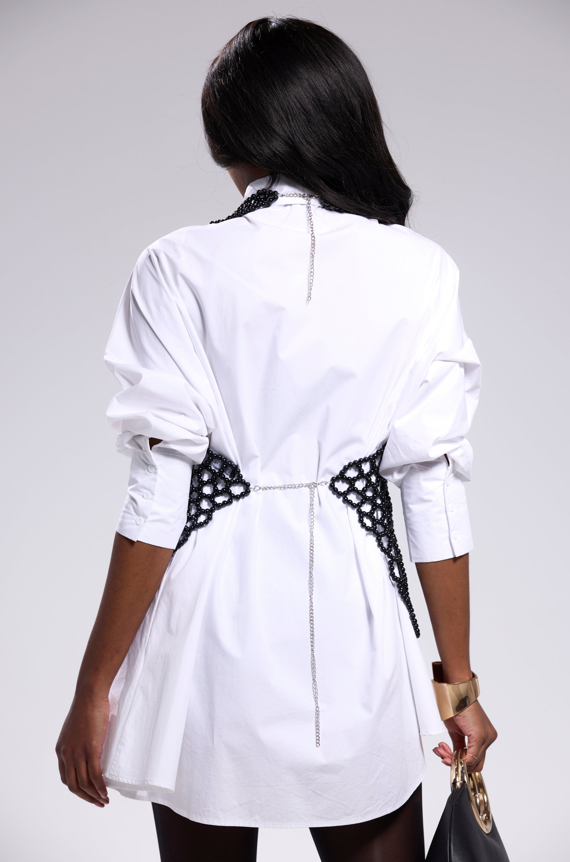 A woman with long dark hair stands facing away, wearing a white shirt-dress with black crochet detail, a chain belt, gold cuff bracelet, black clutch purse, and the GETTIN HOT IN HERE BEADED TOP IN BLACK as a chic accent. 8