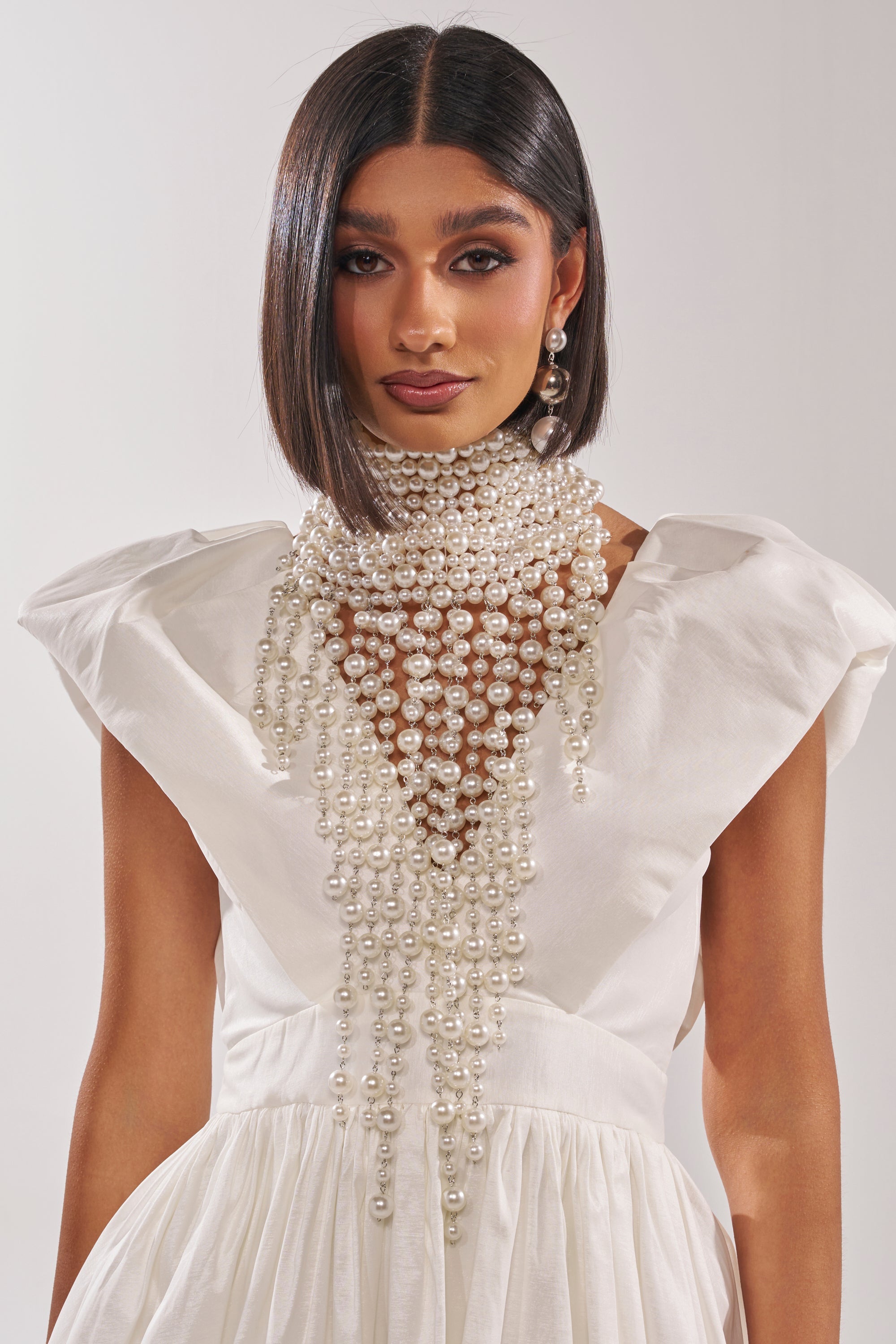 A woman in a white dress with structured shoulders wears the POLLY PEARL STATEMENT NECKLACE, its dramatic pearls cascading over her neck and chest. She completes the luxurious look with sleek hair and large pearl earrings against a light background.