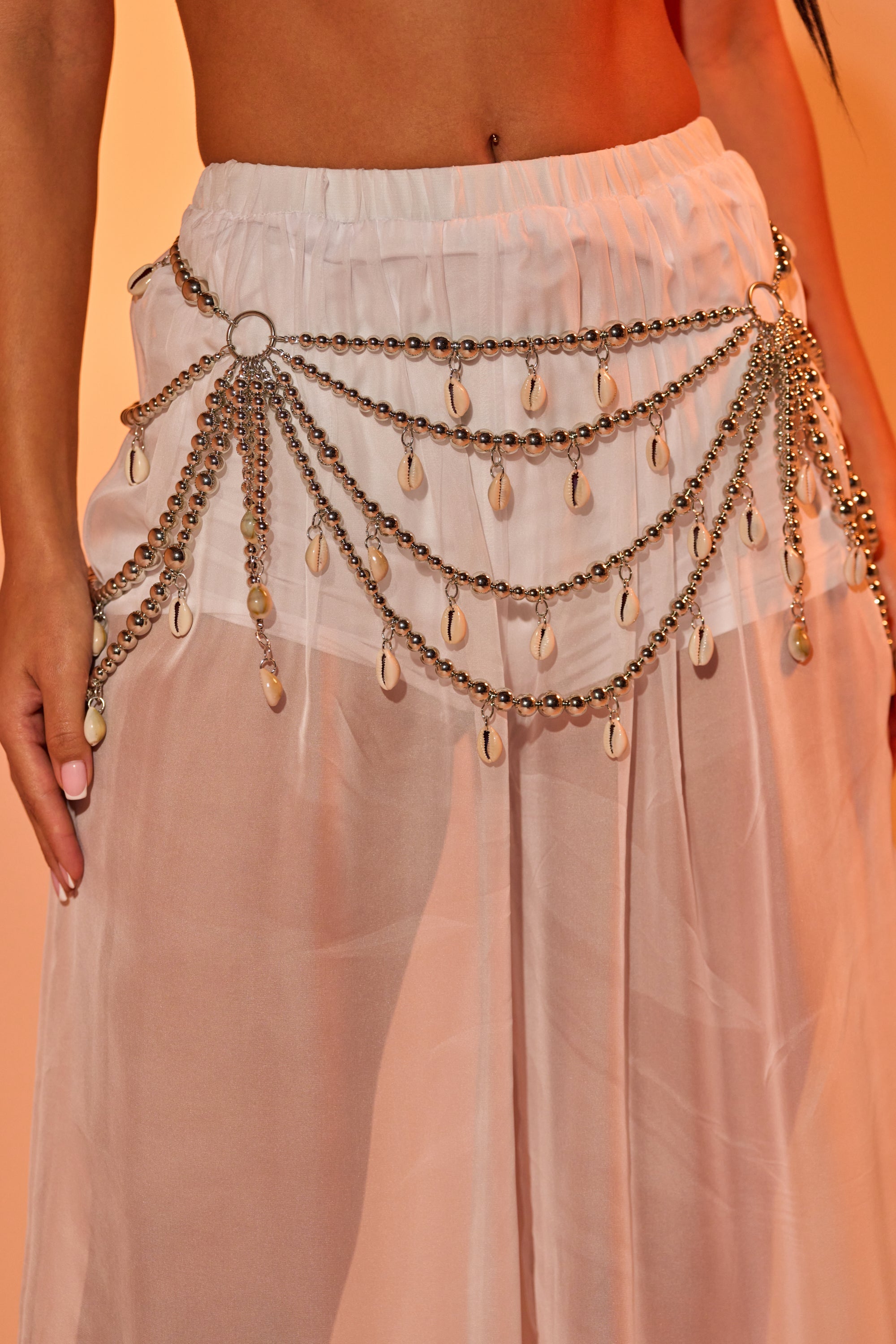 A person styles a sheer, light pink skirt with the TOO CHIC SEASHELL BELT, featuring multiple strands and dangling shell-like charms for a chic, bohemian festival look. 0