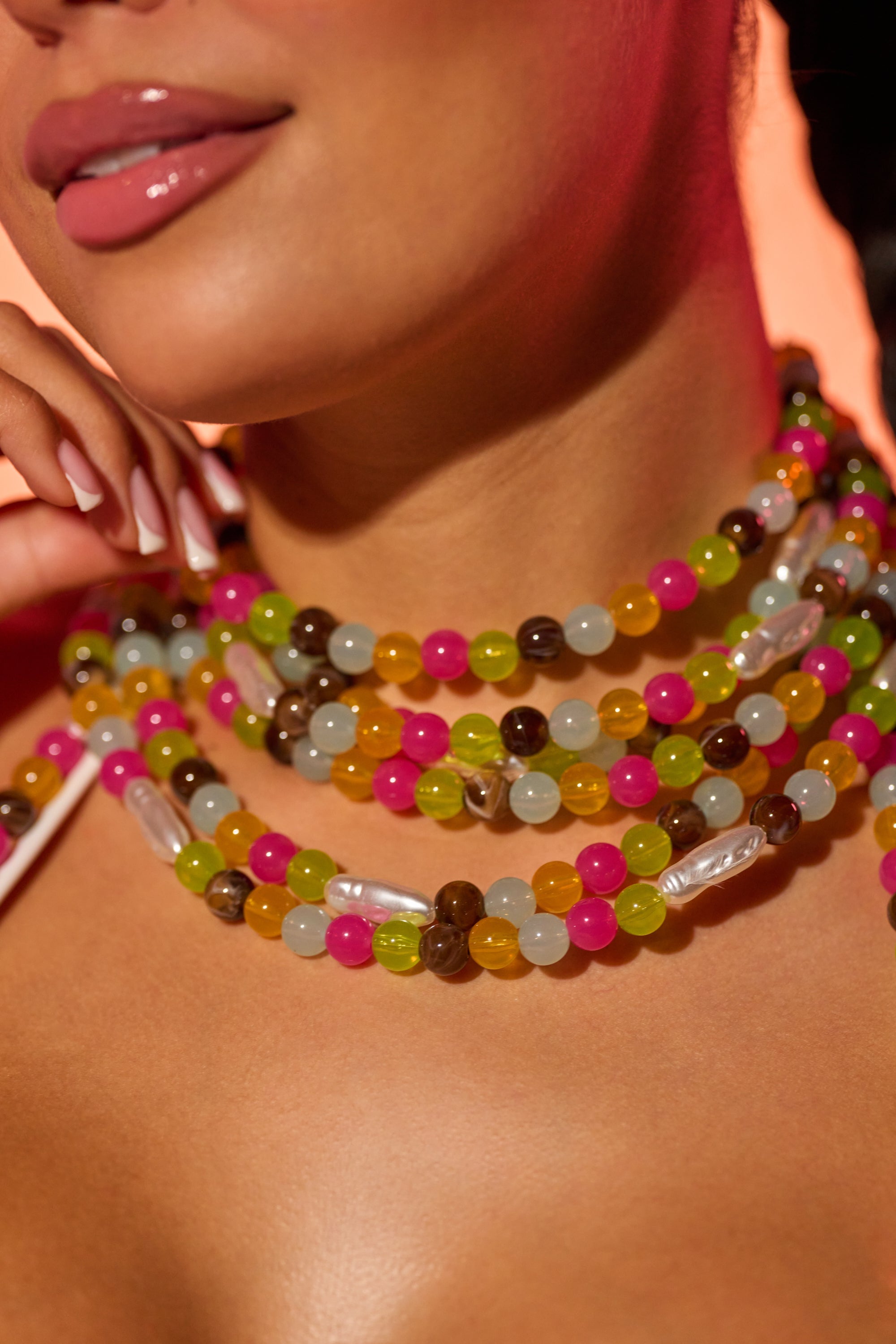 A close-up of a woman wearing glossy pink lipstick and the IN YOUR DREAMS CHOKER, a layered necklace with colorful beads—pink, green, yellow, white, and brown—and faux pearl accents. Her hand gently touches her neck. 0
