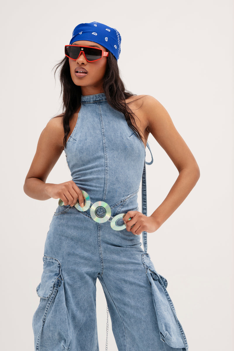 A person confidently poses in a sleeveless denim jumpsuit with cargo pockets, accessorized by the PLAY IT AGAIN CD BELT. They sport a blue bandana, red sunglasses, and hold the belt against a plain, light background.