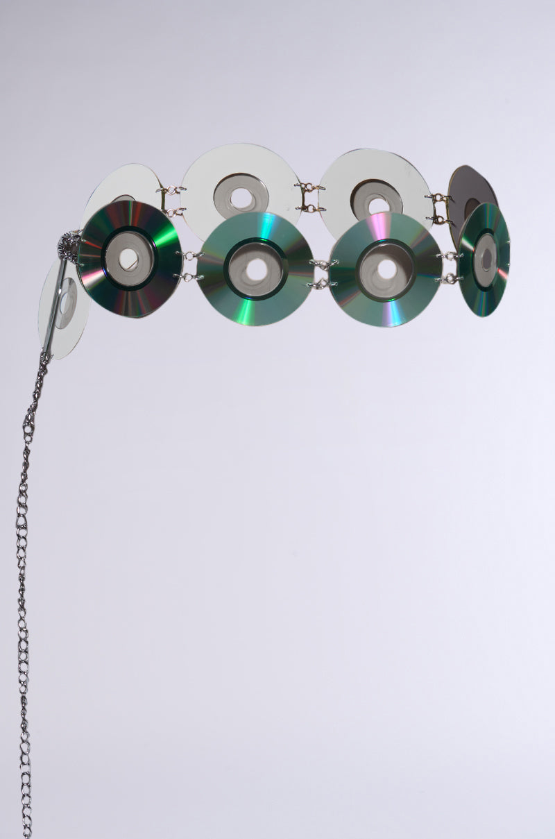 The PLAY IT AGAIN CD BELT is a Y2K accessory, crafted from holographic CD discs with metallic rings forming circular patterns. It features a chain elegantly dangling from one end against a plain, light gray background.