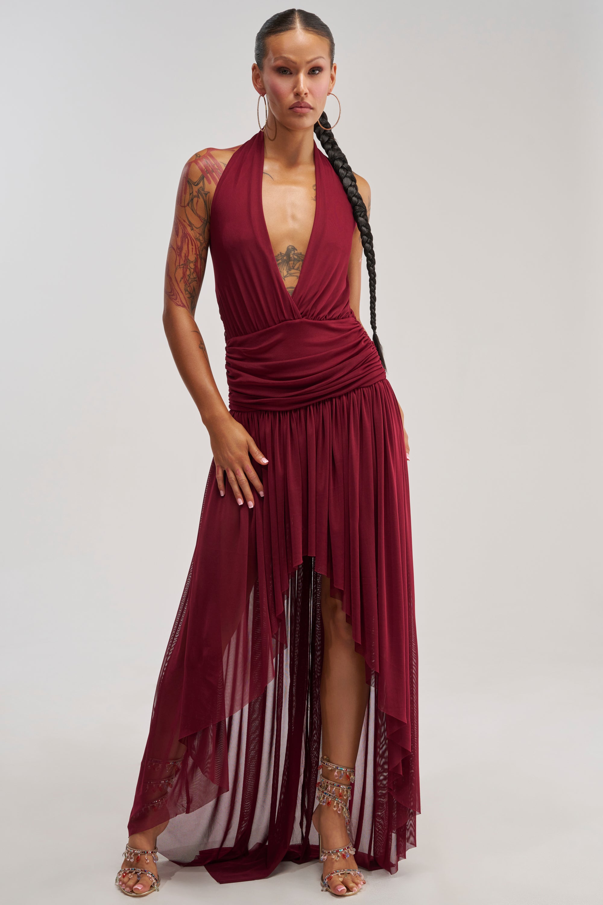 A woman stands against a light gray background wearing the LIKE MY STYLE HIGH LOW MIDI DRESS IN BURGUNDY—a sleeveless halter dress with an asymmetrical hem—paired with gold high-heeled sandals, large hoop earrings, and a long braided ponytail.