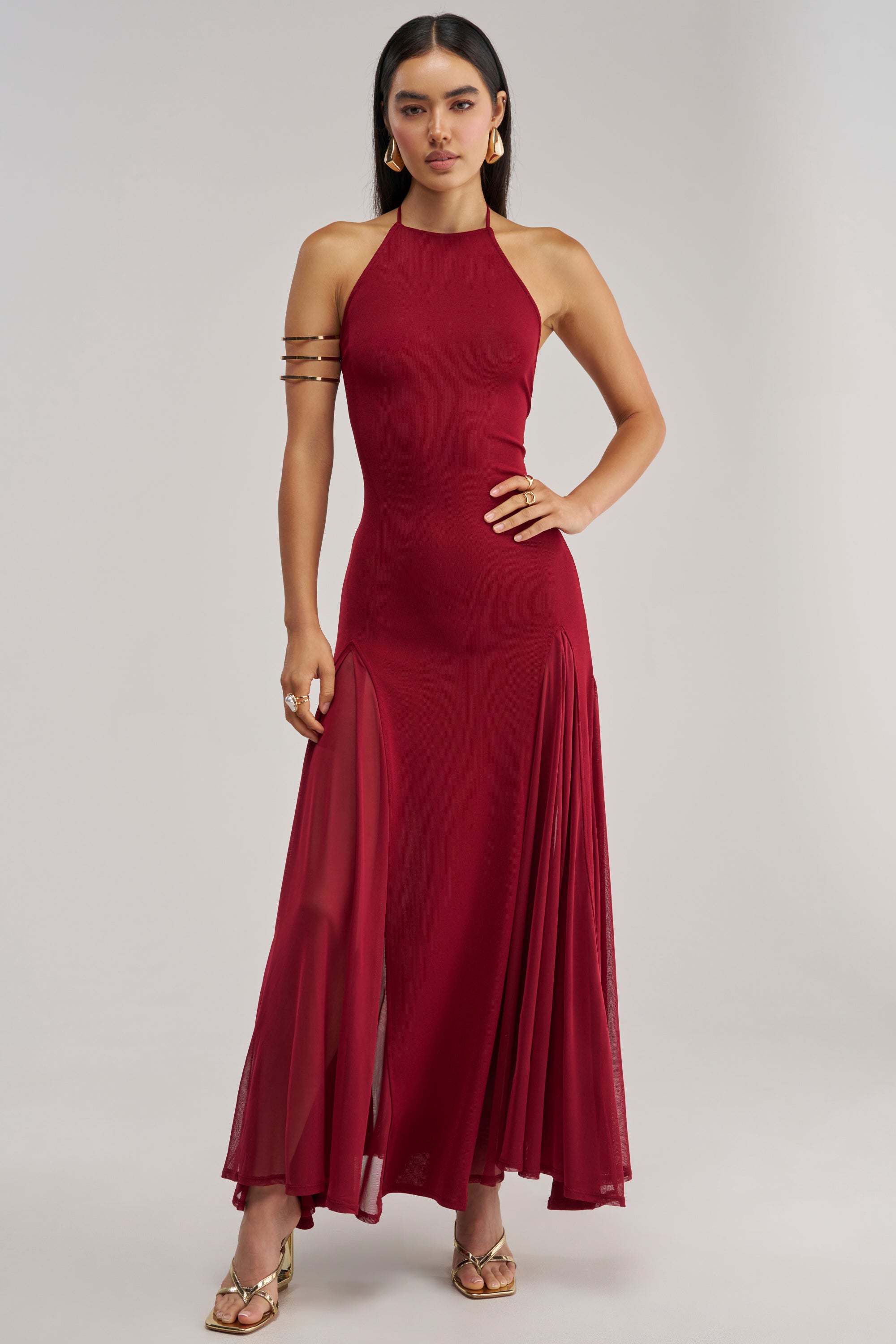 A woman stands with one hand on her hip against a plain background, wearing the stunning CATCH ME DANCING HIGH NECK MAXI DRESS—a sleeveless, floor-length burgundy maxi dress featuring halter neckline and sheer skirt panels—accented by gold earrings and sandals; the perfect special occasion look for a wedding guest.