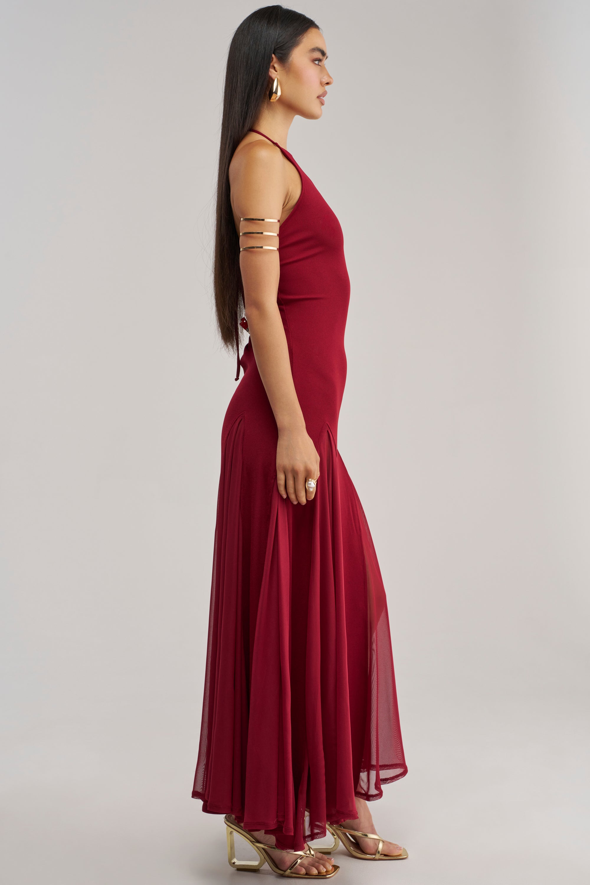 A woman stands in profile against a plain background, wearing the floor-length CATCH ME DANCING HIGH NECK MAXI DRESS—a burgundy halter neck style with a mesh hem—offering an elegant look ideal for wedding guests or special occasions. She accessorizes with a gold arm cuff on her upper arm, gold hoop earrings, a statement ring, and strappy gold heeled sandals.