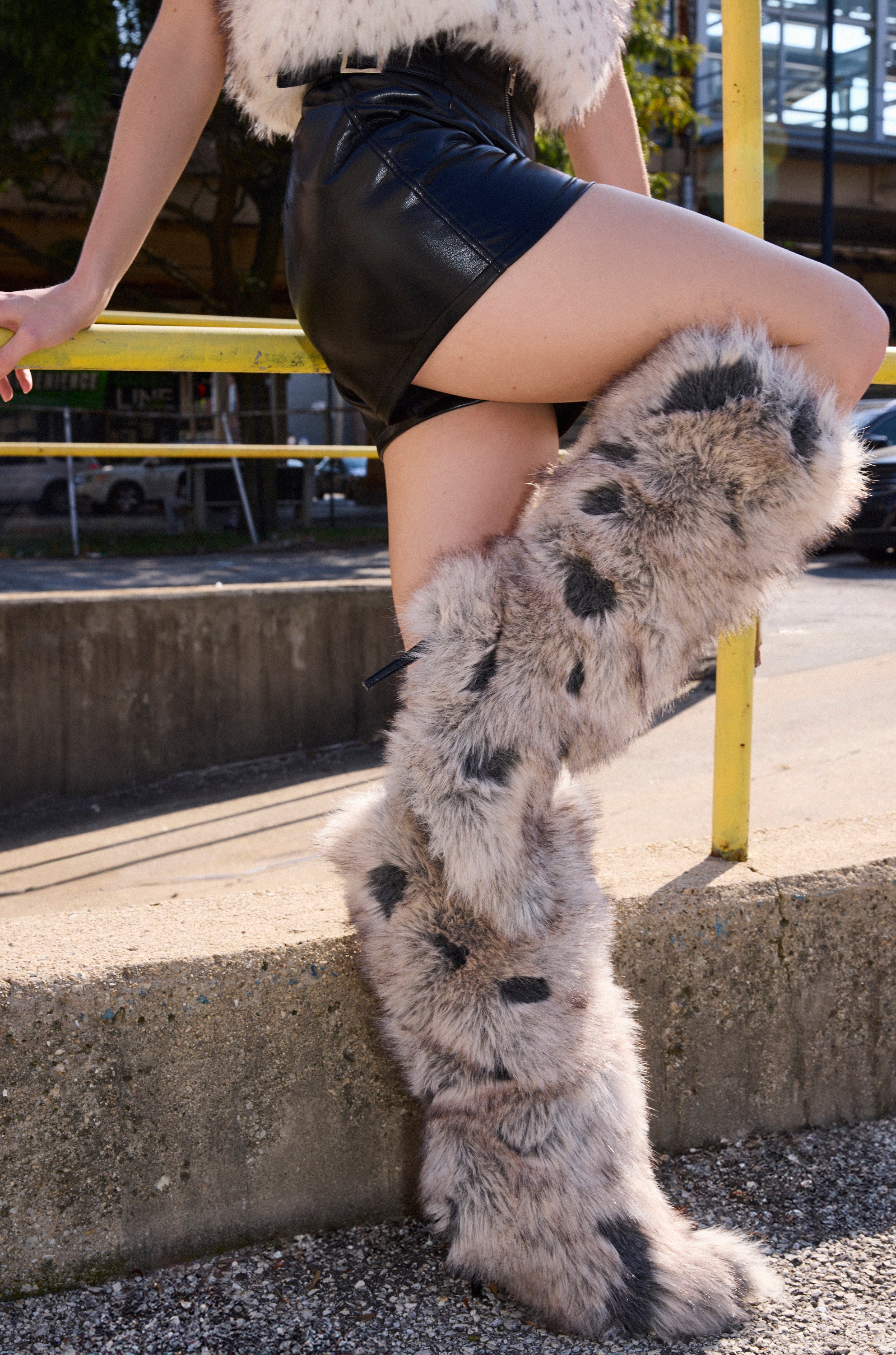 Wearing a faux fur vest, black leather skirt, and AZALEA WANG DEBBY GREY MULTI FUR STILETTO BOOTIE, a person leans against a yellow railing outdoors with one knee bent as sunlight creates dramatic shadows.