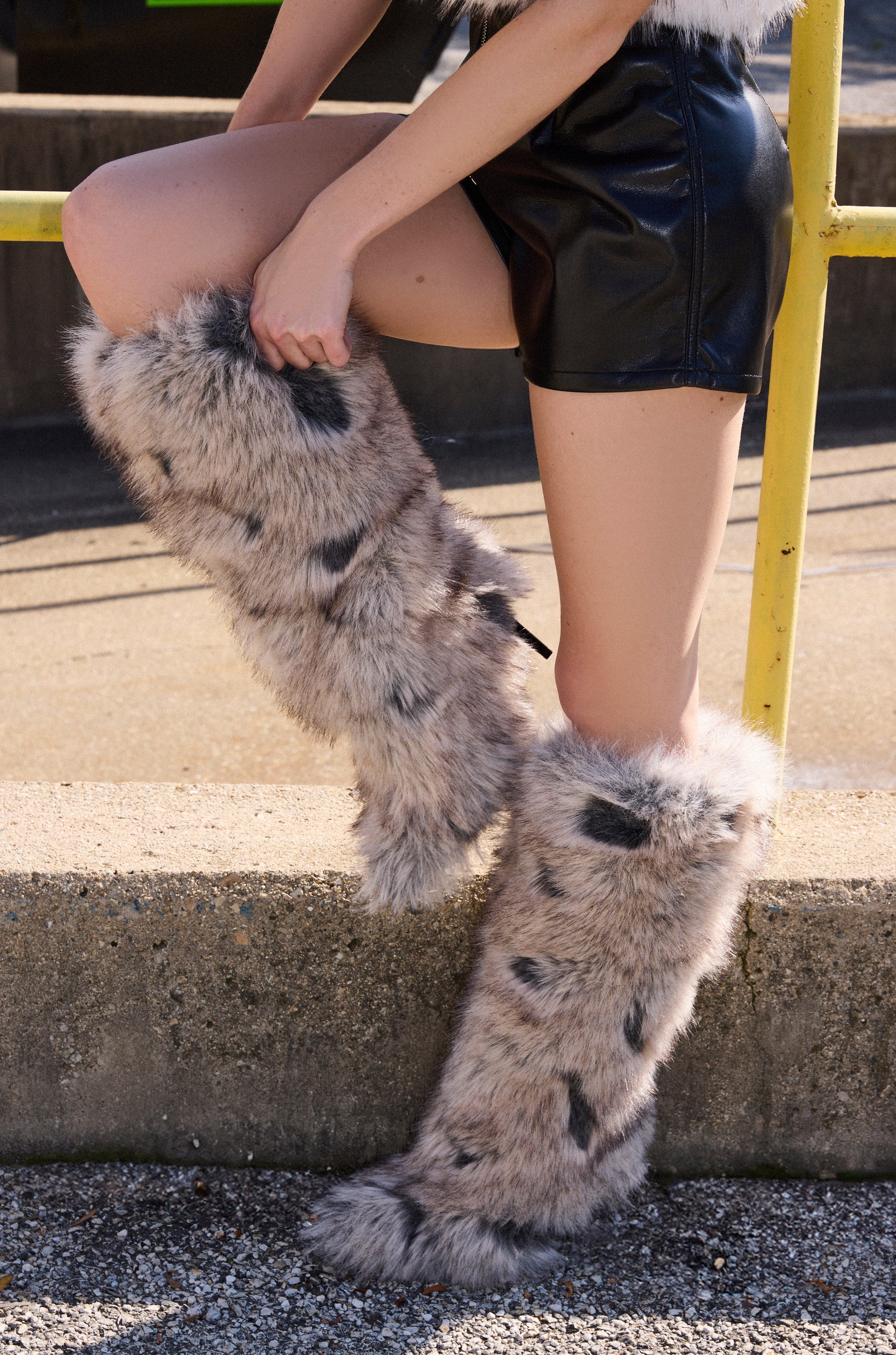 Wearing black leather shorts and furry leg warmers, this person stands outdoors by a concrete ledge. The AZALEA WANG DEBBY GREY MULTI FUR STILETTO BOOTIE peeks out beneath the leg warmers as their hands rest on one knee.