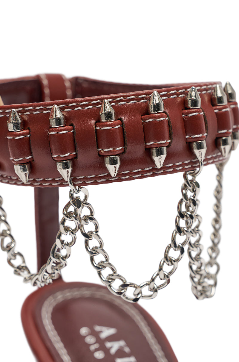 A close-up of the AKIRA GOLD DELCROIX BROWN STILETTO SANDAL shows a brown faux leather strap with silver spikes, white stitching, hanging silver chains, and part of the brand name visible on the insole. 7