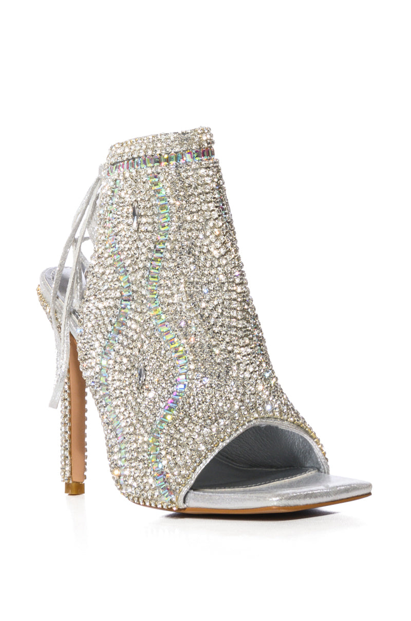 The AZALEA WANG DELANIE SILVER STILETTO SANDAL is a stunning silver shoe with rhinestones in a wavy design. It features a peep toe, open back, lace-up details, and is set on a slender heel for an elegant and charming look.