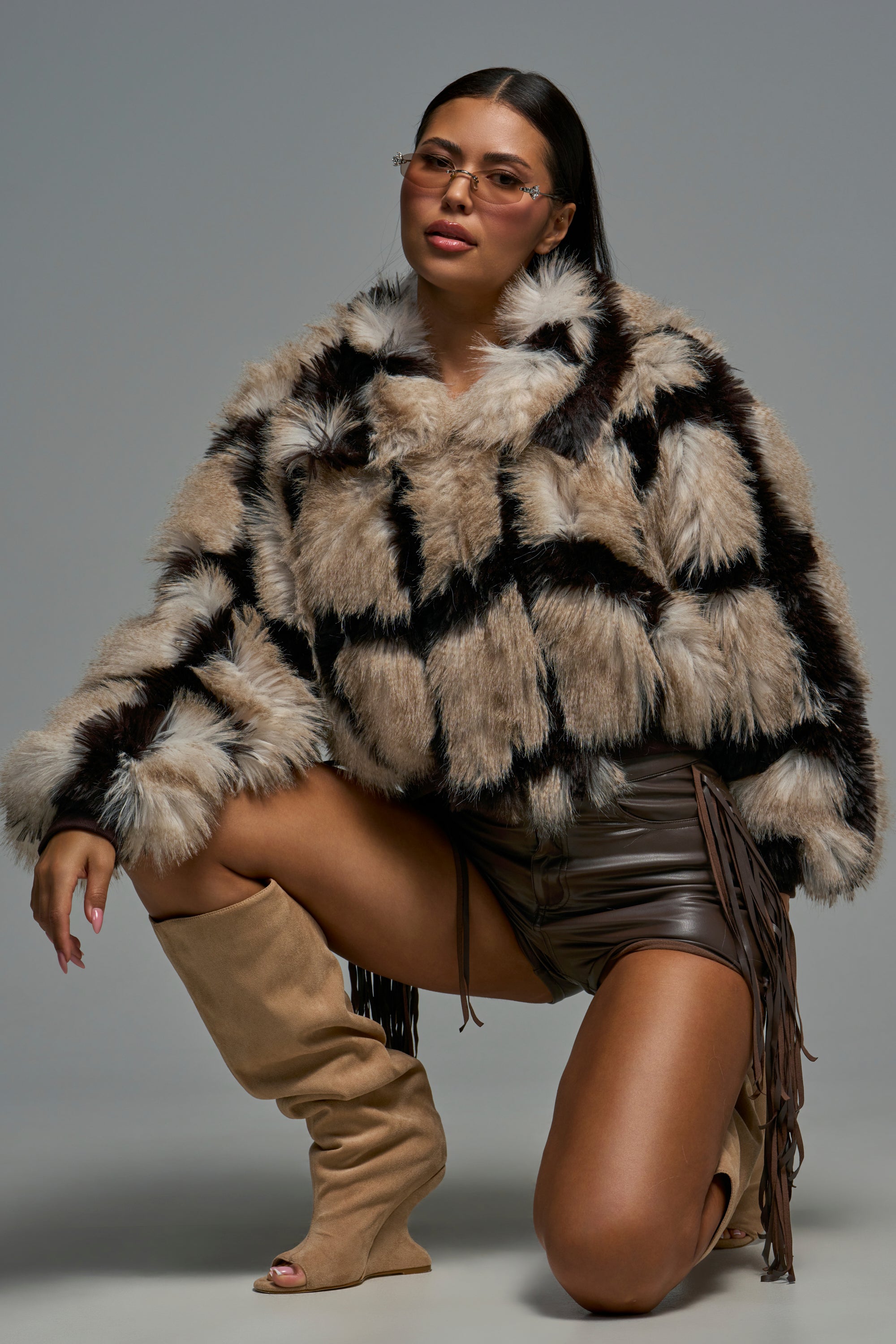 Wearing the DELILAH FAUX FUR BOMBER, a woman in tinted glasses pairs her brown leather shorts with tan knee-high boots for a chic winter look. Kneeling on one knee against a plain gray background, she gazes confidently at the camera.