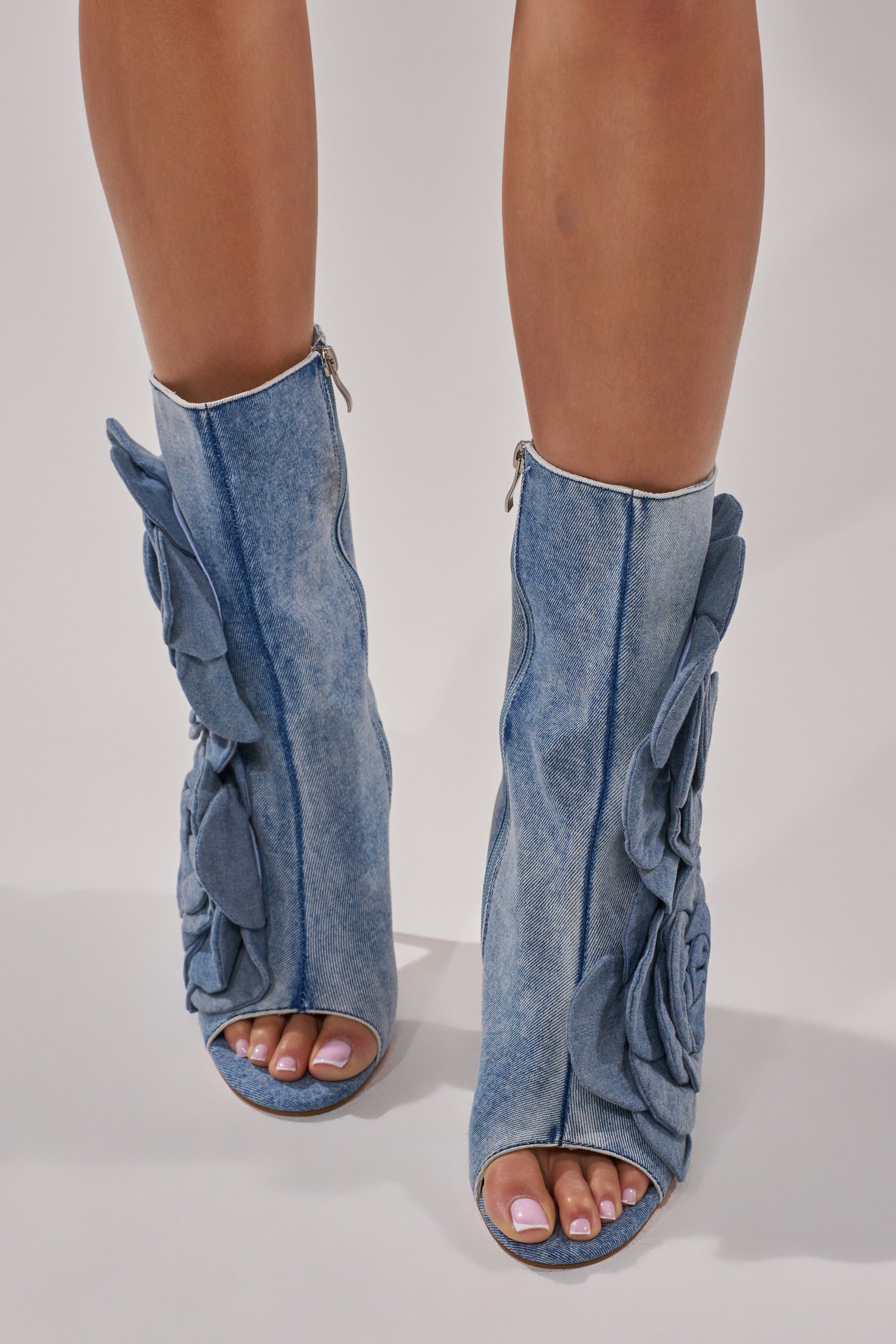A person models the AZALEA WANG DELMARE DENIM FLORAL OPEN TOE BOOTIE—mid-calf booties with large denim flower accents, open toes, and a chic stiletto heel—while standing on a light surface.
