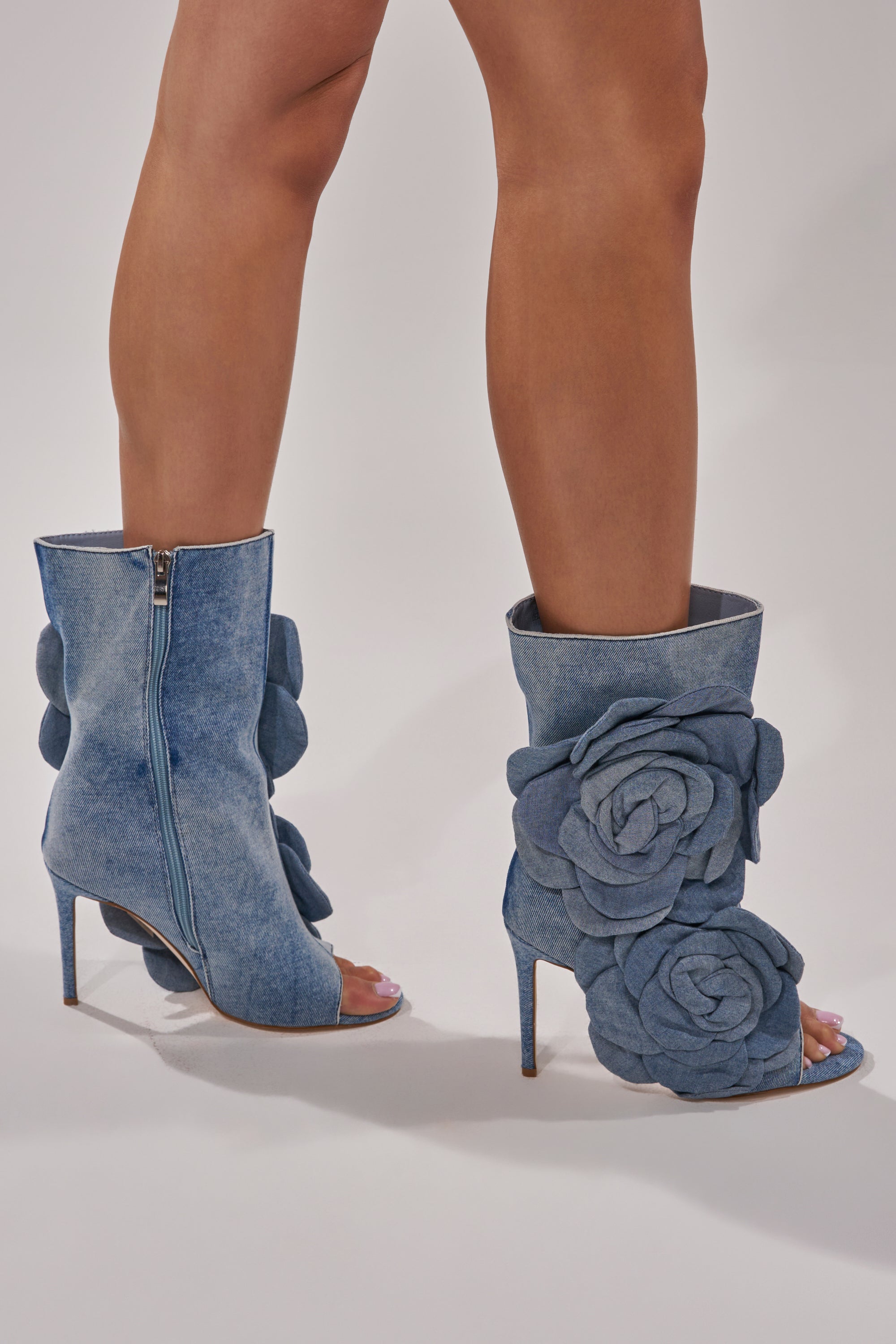 A person stands confidently on a white background wearing the AZALEA WANG DELMARE DENIM FLORAL OPEN TOE BOOTIE, crafted from denim and embellished with large denim flower appliqués.