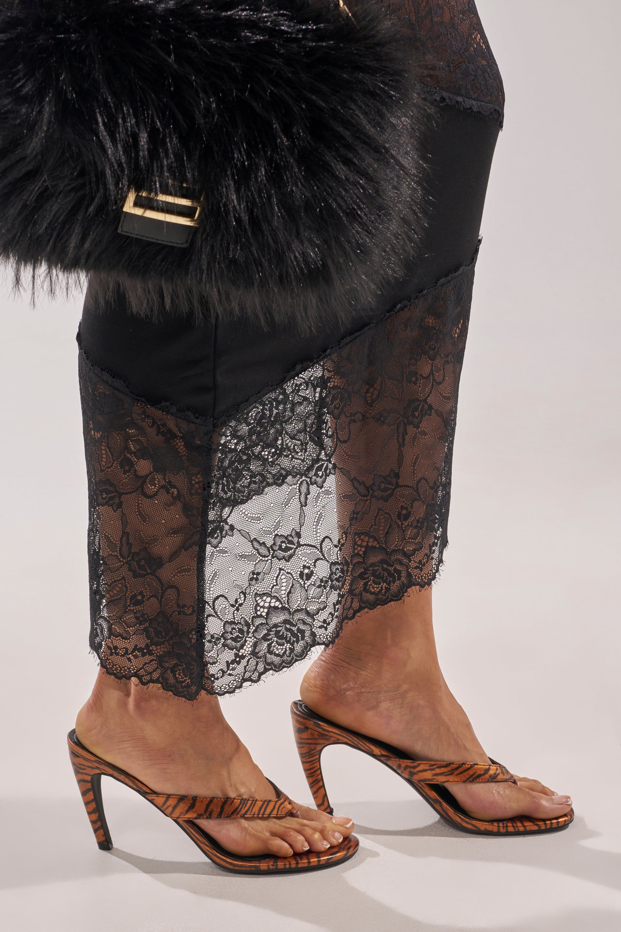 A person in a black lace skirt, holding a fluffy black handbag, and wearing AKIRA GOLD DELYSE ORANGE TIGER PRINT PU SLIP ON STILETTO SANDALS with unique curved heels and comfort cushion insoles. 2
