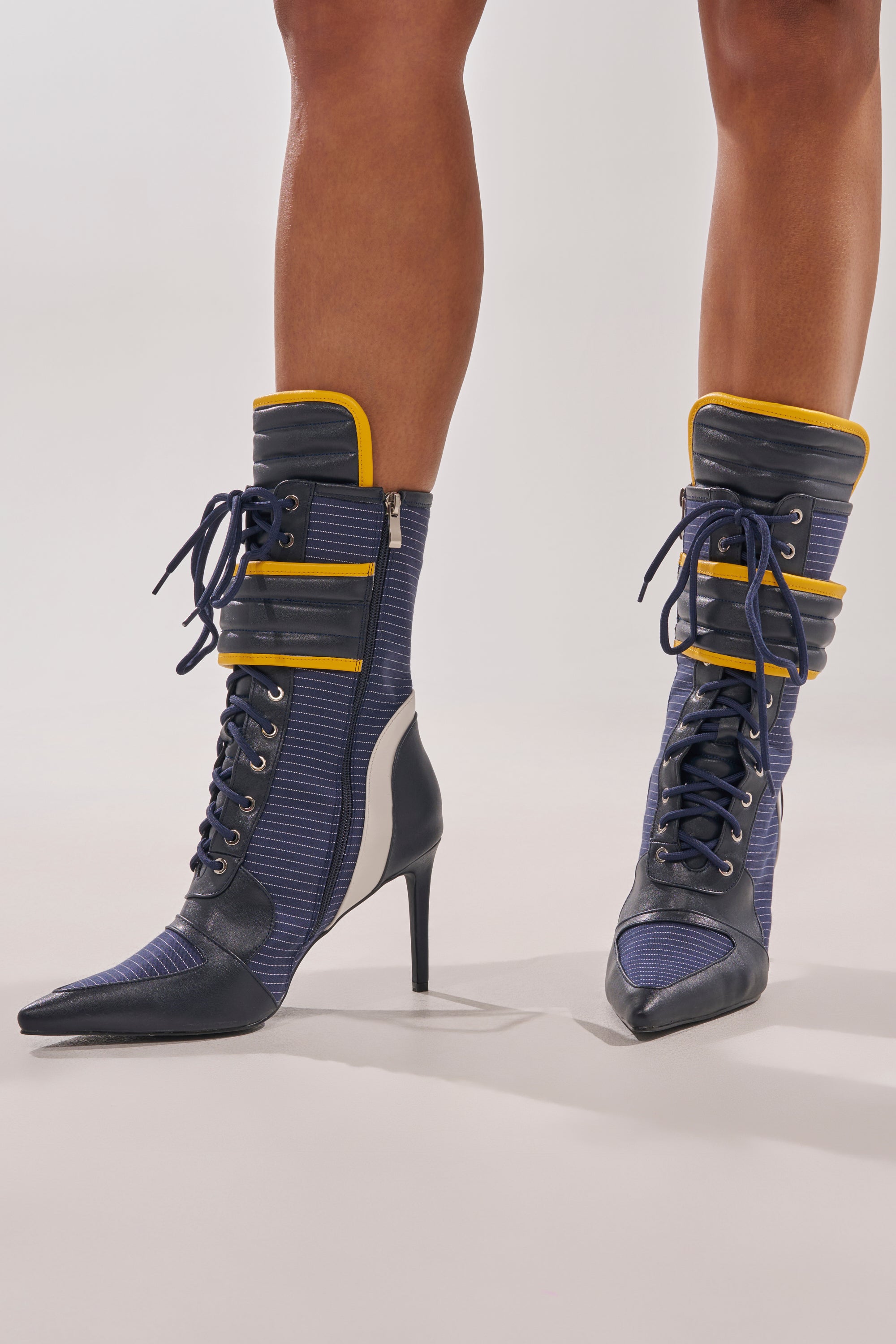 A person wears the AKIRA GOLD DEMAND NAVY STILETTO BOOTIE, featuring pointed toes, high heels, lace-up design, and a blue and gold color scheme with sporty details, standing on a light background. 0