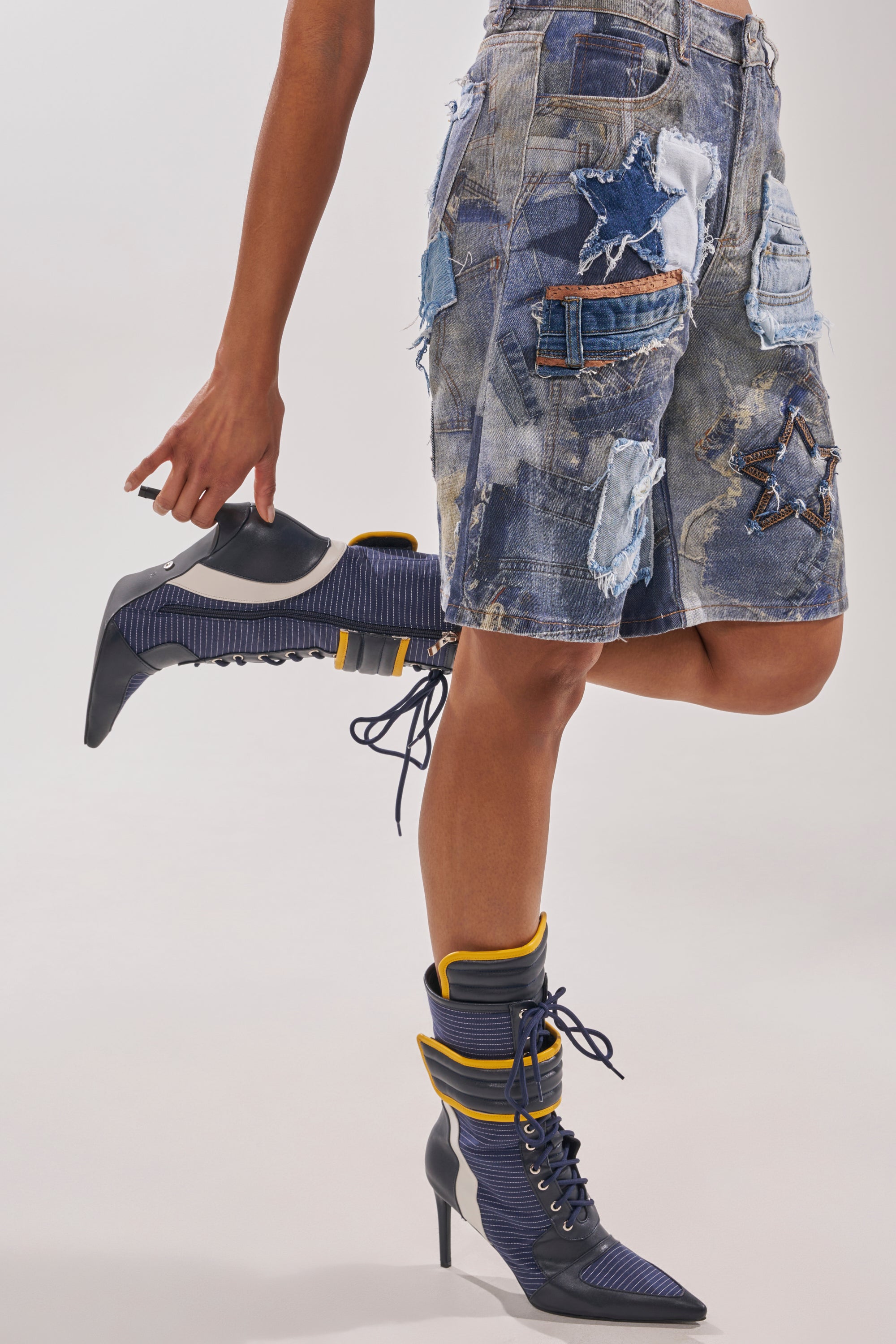 A person stands on one leg against a plain background, holding an AKIRA GOLD DEMAND NAVY STILETTO BOOTIE while wearing a patchwork denim skirt and the matching blue pointed toe booties with yellow trim. 1