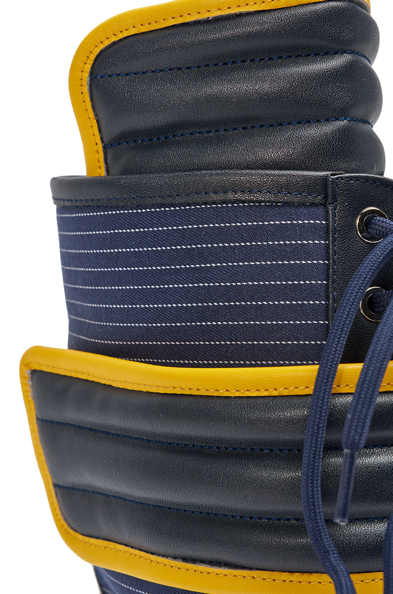 Close-up of the AKIRA GOLD DEMAND NAVY STILETTO BOOTIE, featuring padded navy blue faux leather, yellow trim, horizontal white stitching, and partially visible blue laces on the right. 5