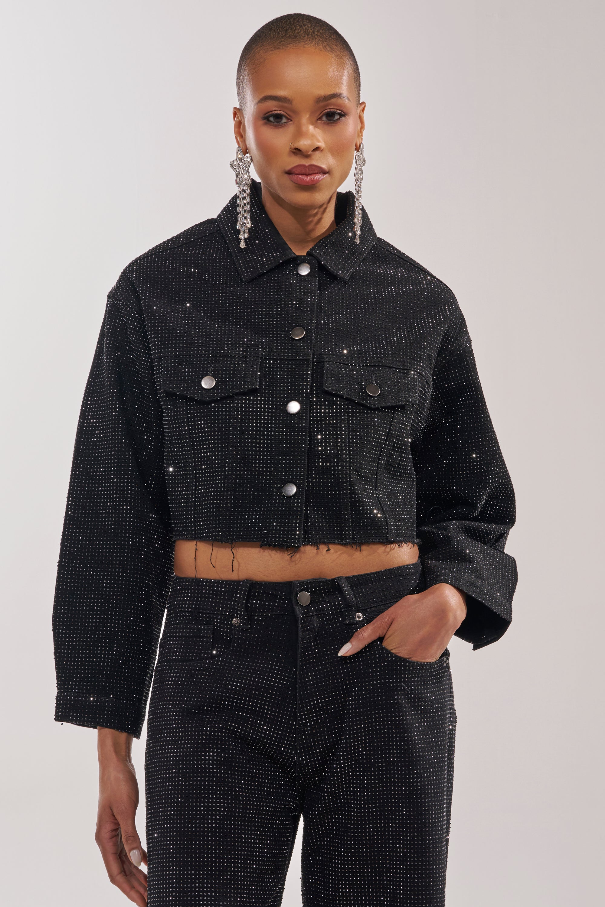 Wearing the DEMI EMBELLISHED DENIM JACKET IN BLACK and matching rhinestone high-waisted pants, a person with short hair and long beaded earrings poses against a plain background, showcasing bold statement streetwear style.