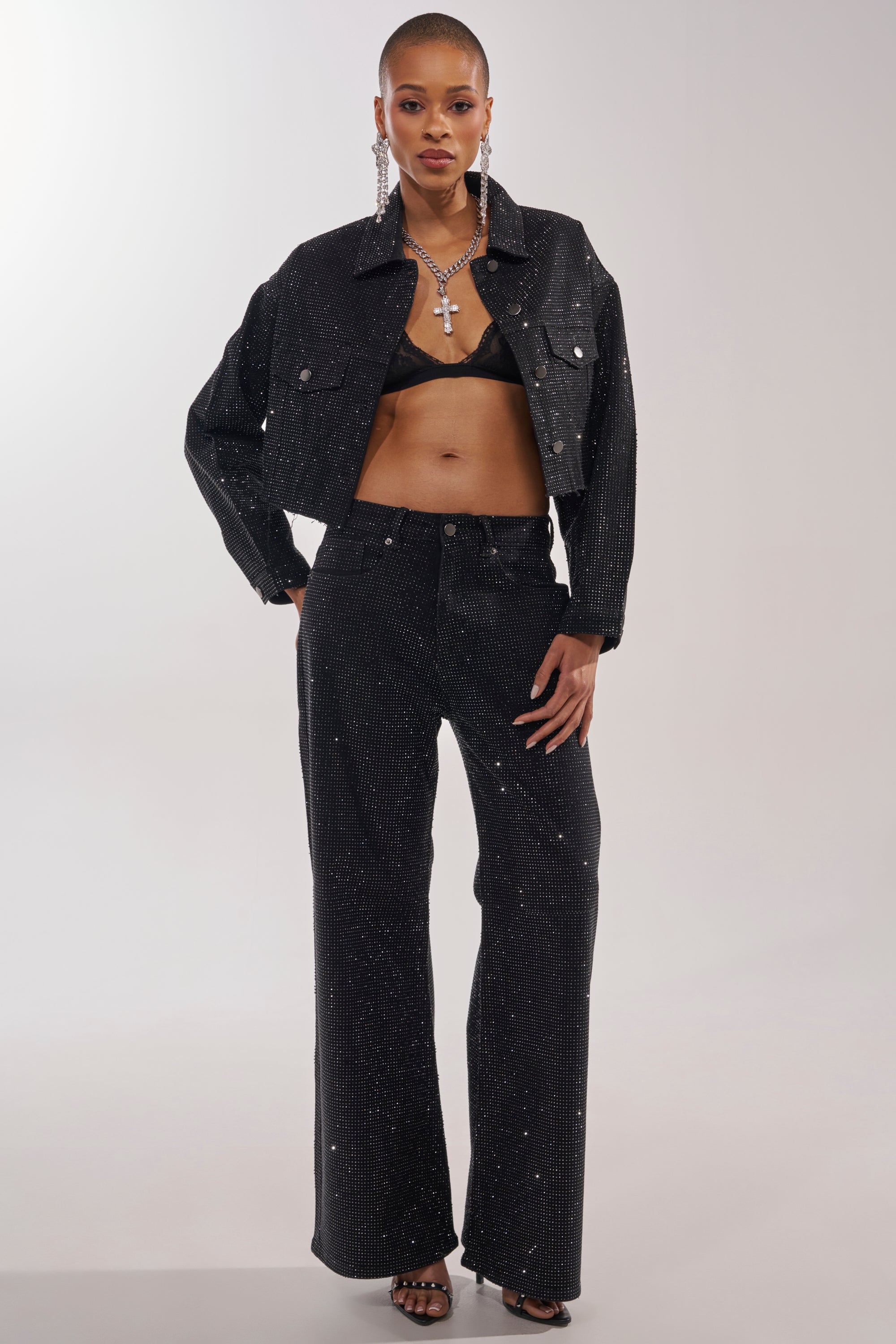A woman stands confidently with one hand on her hip, wearing sparkly black cropped jacket, black bra, hoop earrings, strappy black heels, and the DEMI EMBELLISHED RELAXED DENIM IN BLACK. She completes the look with bold makeup. 3
