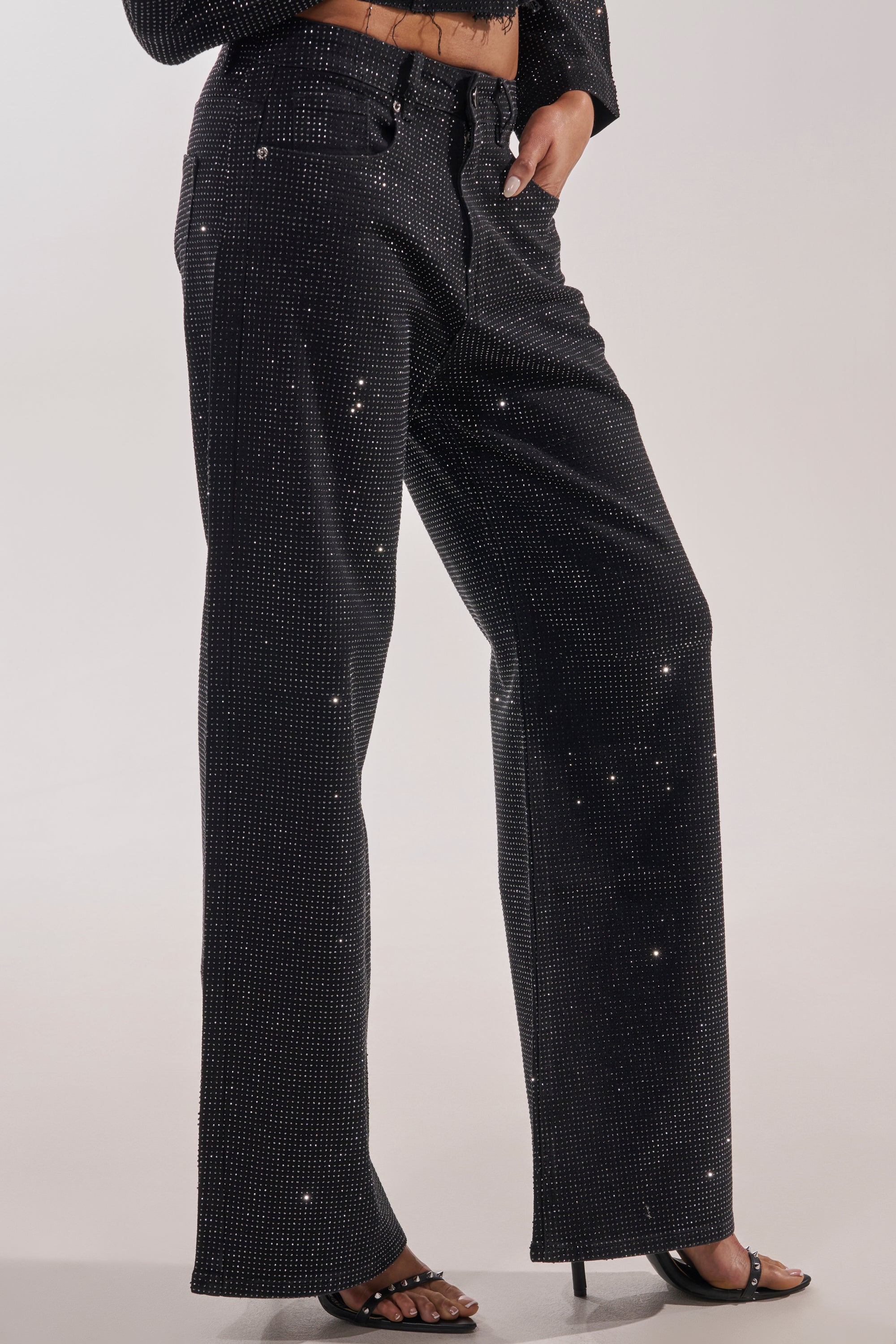 A person models the DEMI EMBELLISHED RELAXED DENIM IN BLACK—high-waisted, wide-leg jeans with rhinestone details—paired with black open-toe heels, shown from waist to feet against a plain backdrop. 0