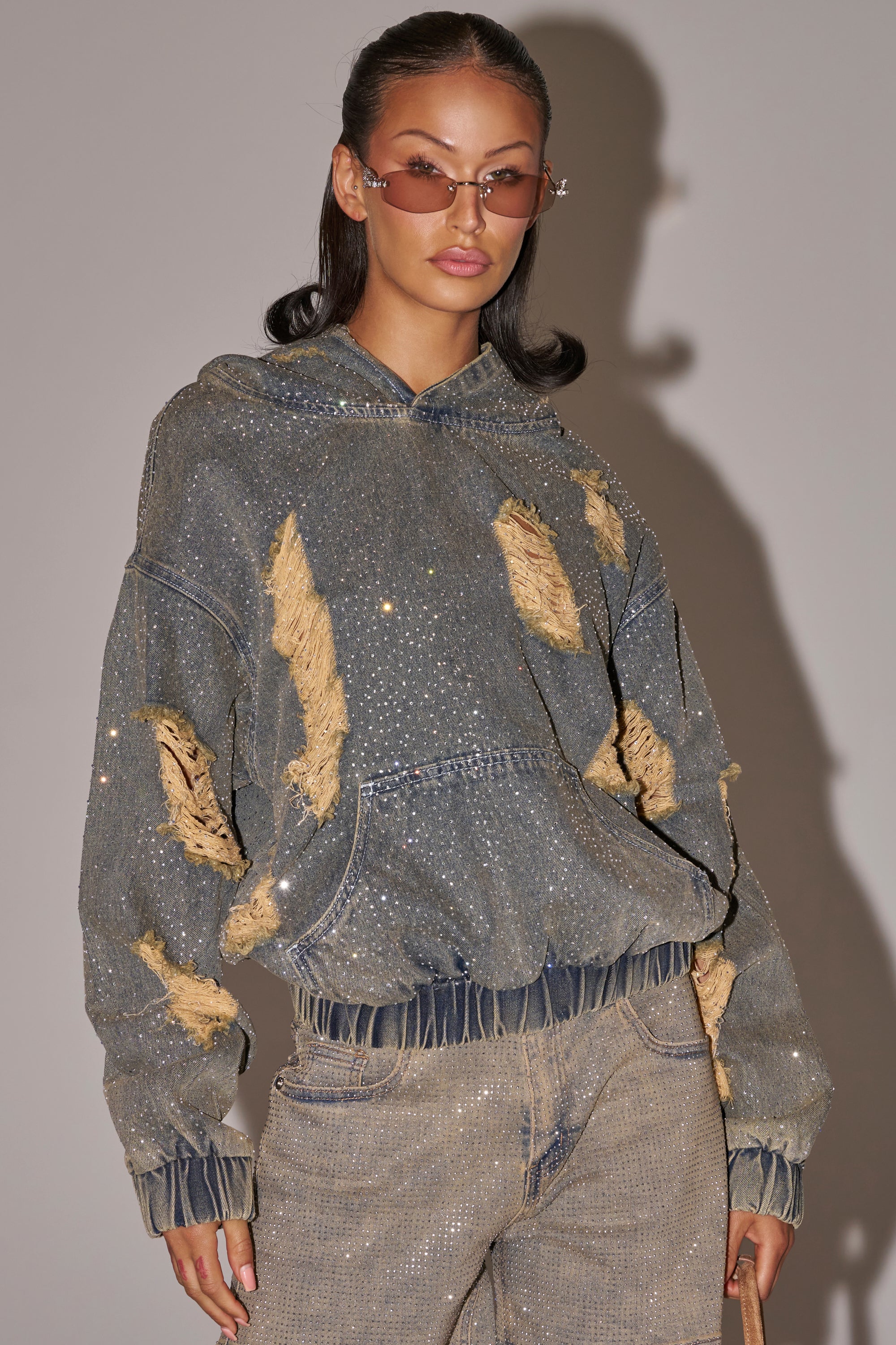 A woman poses in the DEMI RHINESTONE DENIM HOODIE and matching jeans, sporting slicked-back hair, tinted sunglasses, and a small handbag—an ideal festival streetwear look against a neutral backdrop. 1