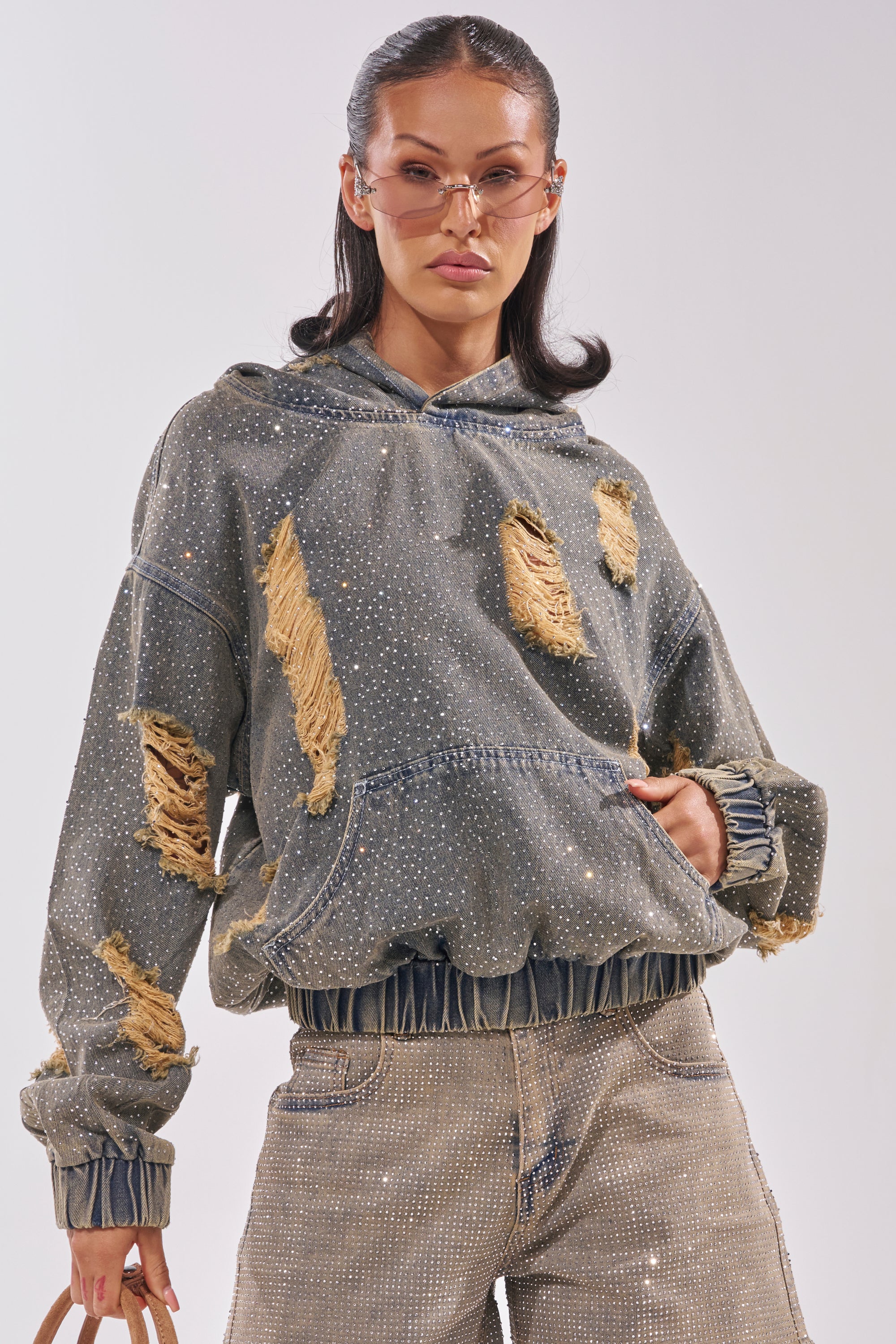 A woman wears the DEMI RHINESTONE DENIM HOODIE with sparkling rhinestones and frayed patches, paired with matching distressed jeans and sunglasses, giving off festival streetwear vibes as she poses with one hand in her pocket. 2